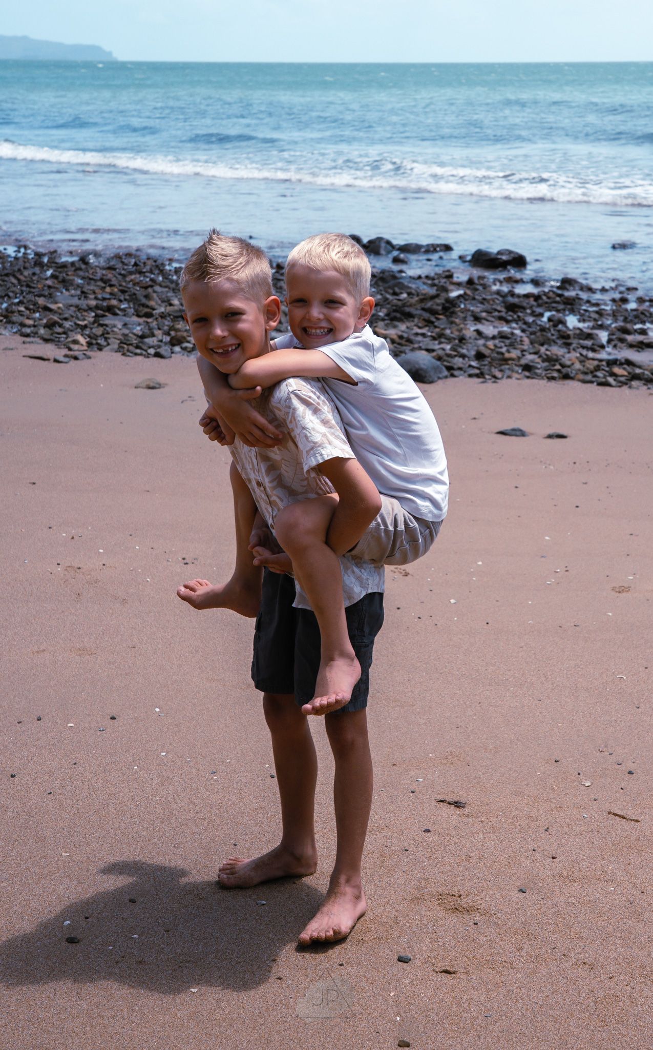 Two children playing together on a sandy beach with ocean waves and rocky shore in the background.