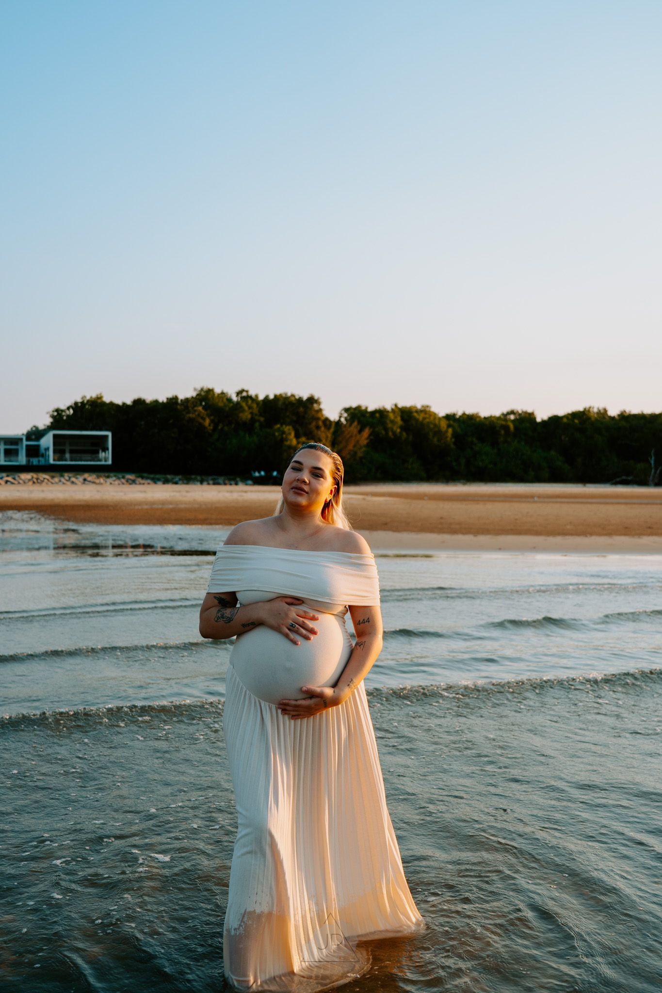 A person in an off-shoulder white dress poses in the ocean waves during golden hour at the beach.