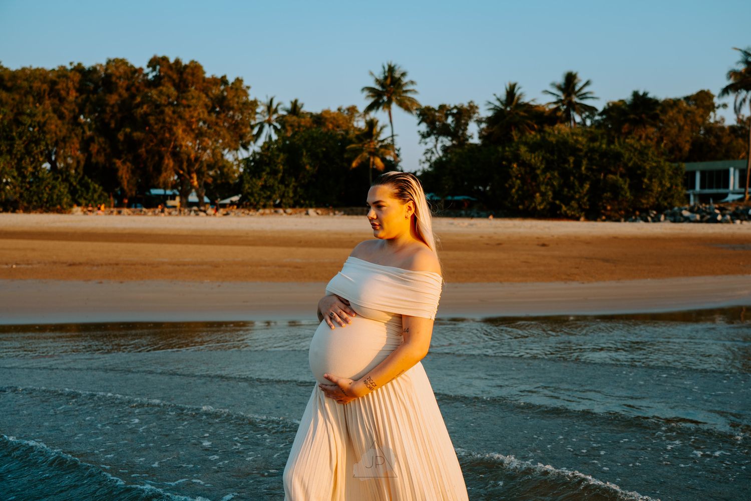 A pregnant woman in a white dress walks along a tropical beach at sunset with palm trees in the background.