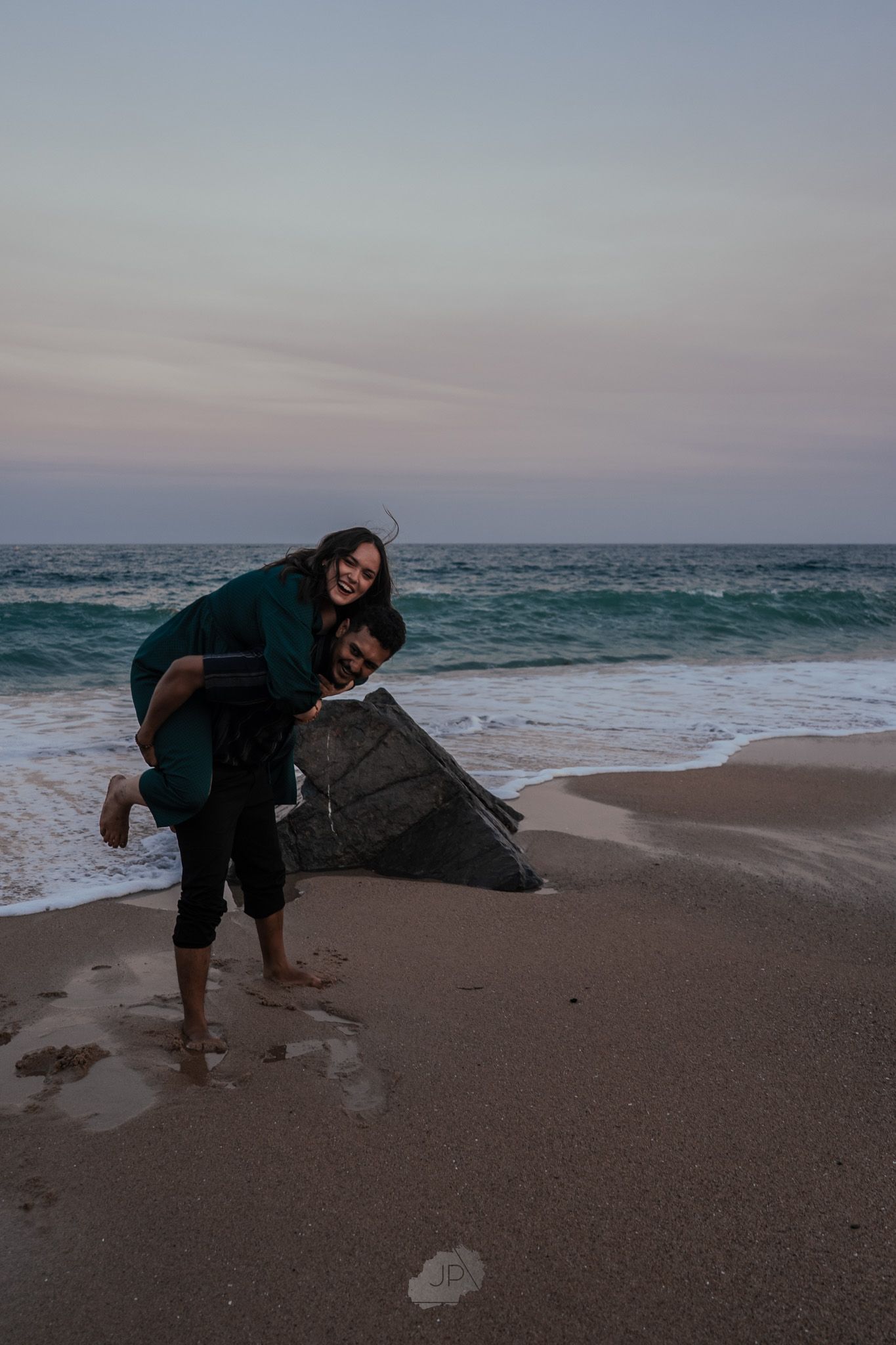 Person in dark clothing bends down to examine seashells on a sandy beach at dusk with ocean waves in the background.