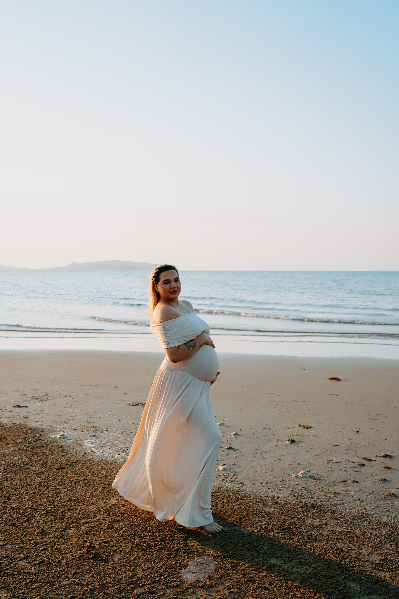 A person in white clothing dances freely on a sandy beach with the calm ocean and clear sky in the background.
