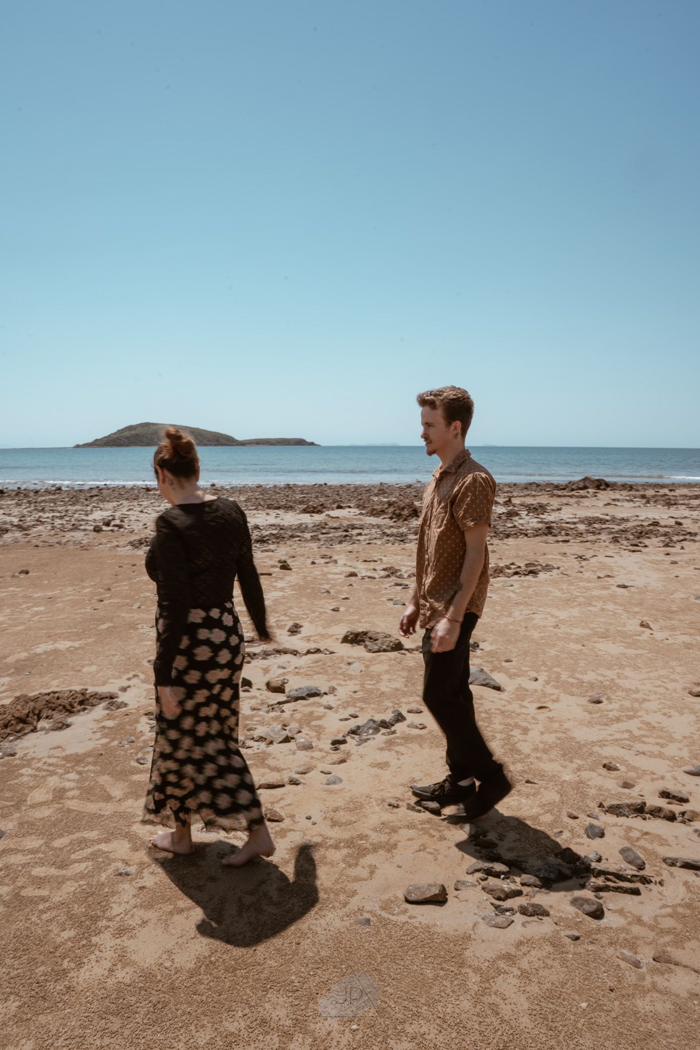 Two people walk across a rocky coastal landscape with clear blue skies and distant hills in view.