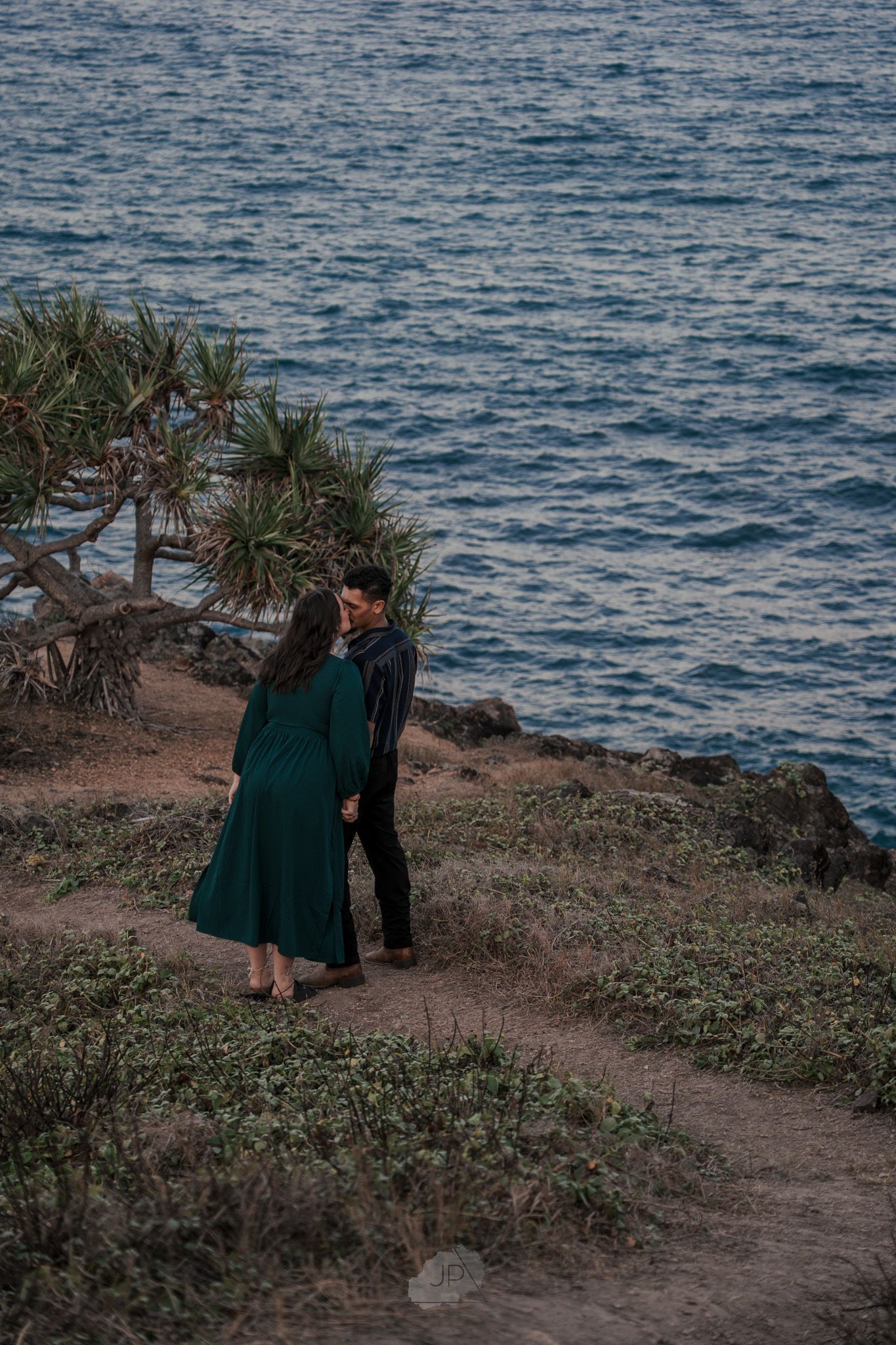 Couple in dark green outfits walking along a scenic coastal path with ocean views at sunset.