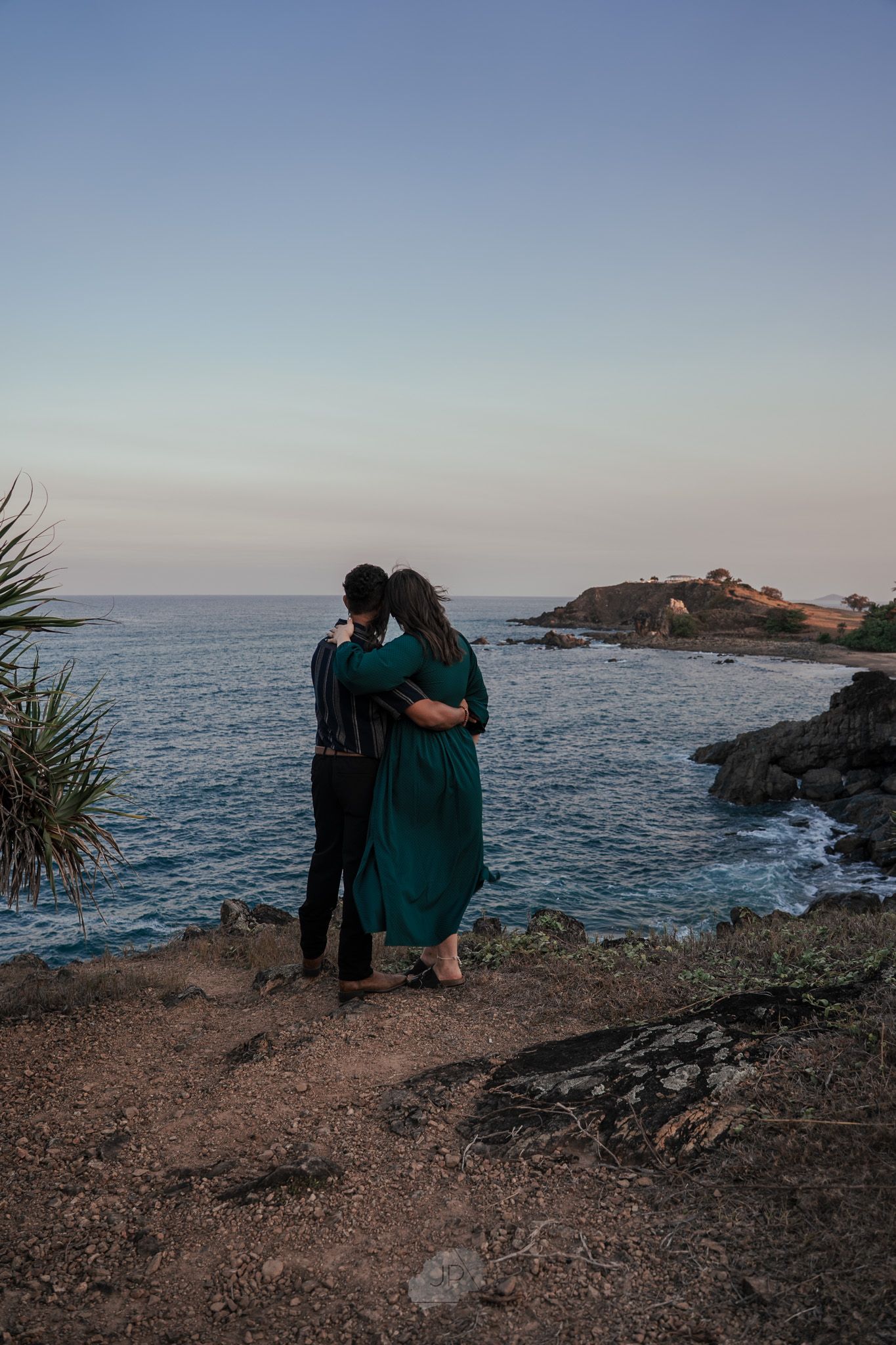 A couple embraces while standing on a rugged oceanside cliff during golden hour.