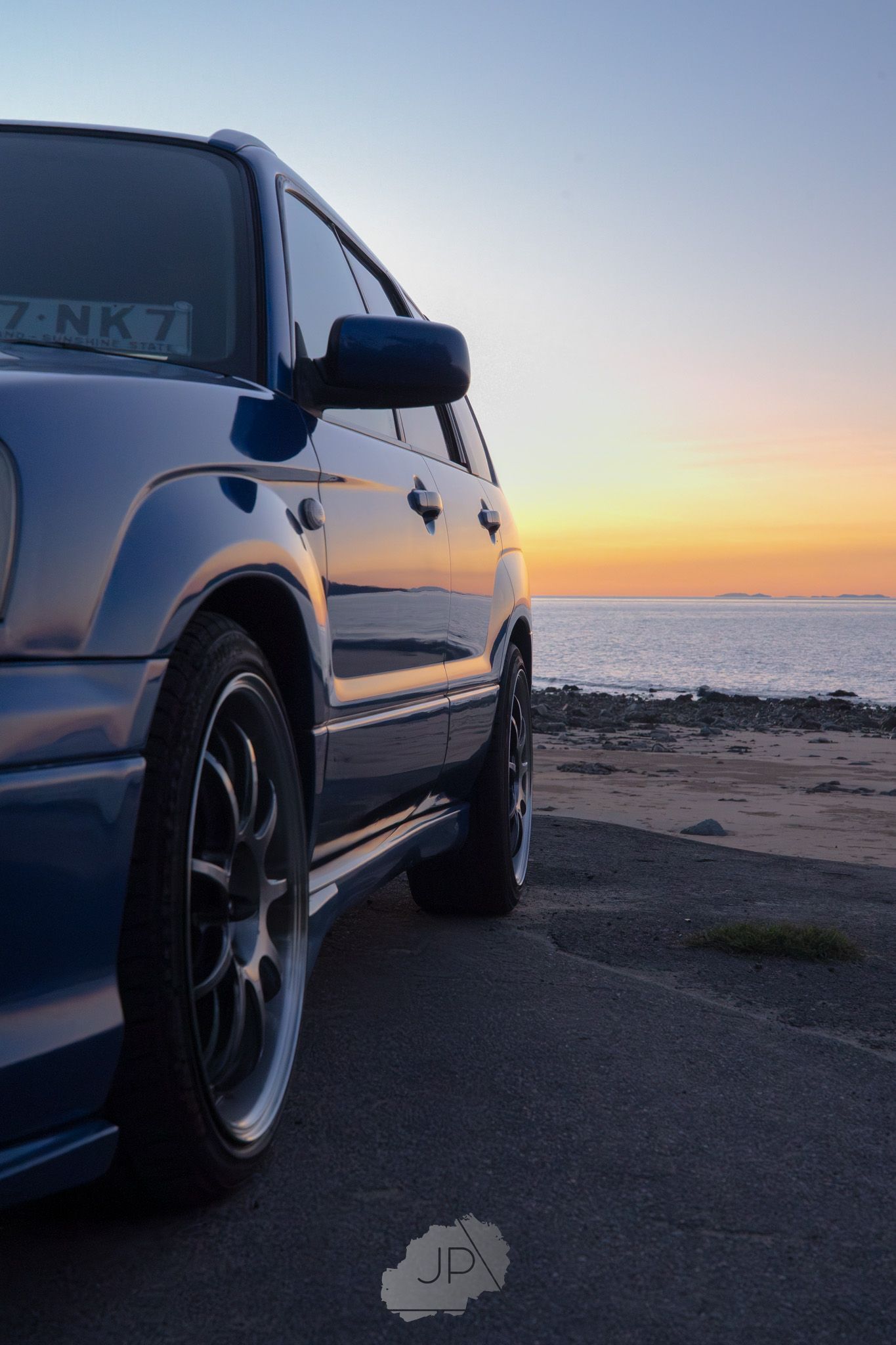 Subaru car parked along a coastal beach at sunset with dramatic silhouettes against the orange sky.