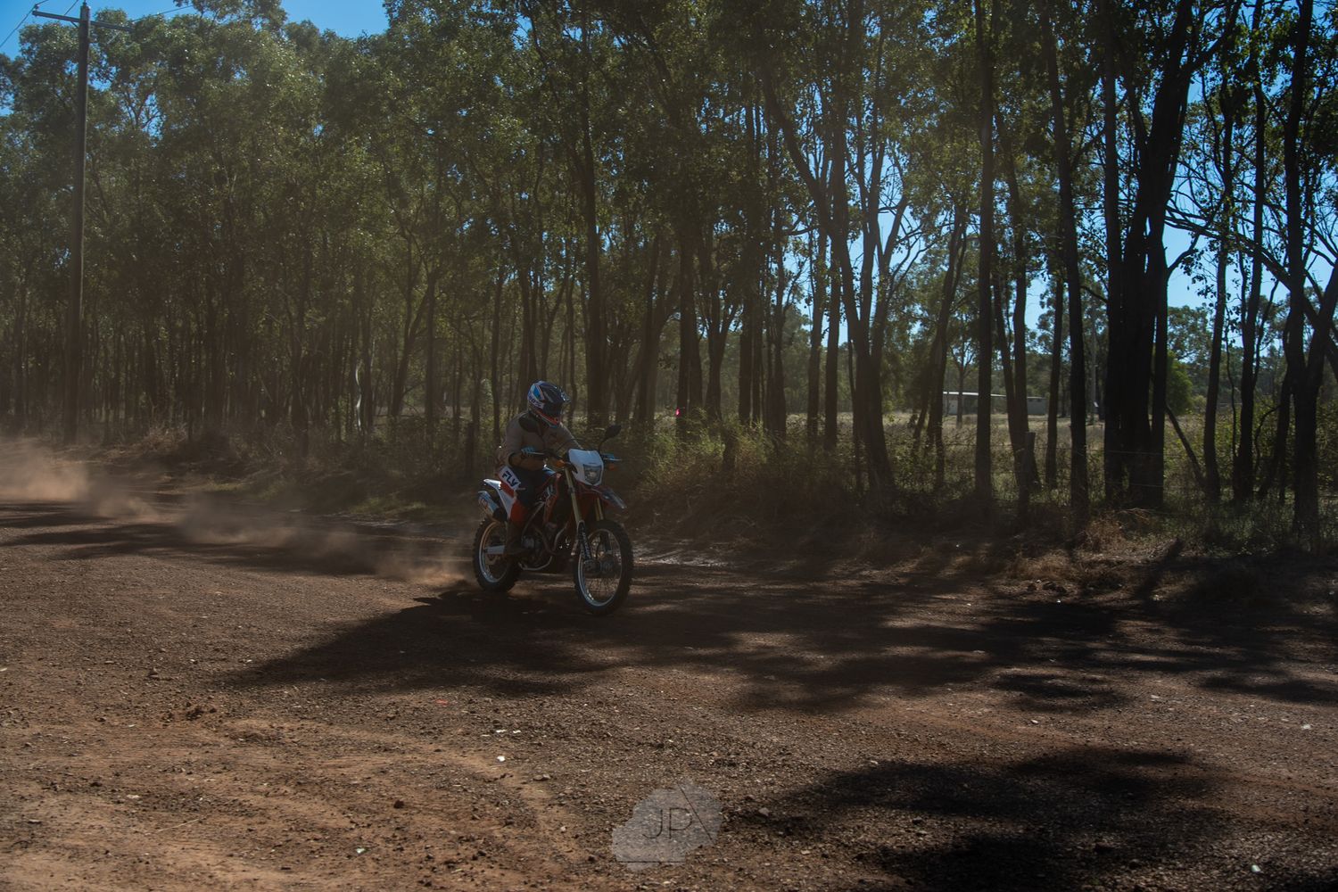 A motorcycle rider navigates a dirt trail through a wooded forest area on a sunny day.