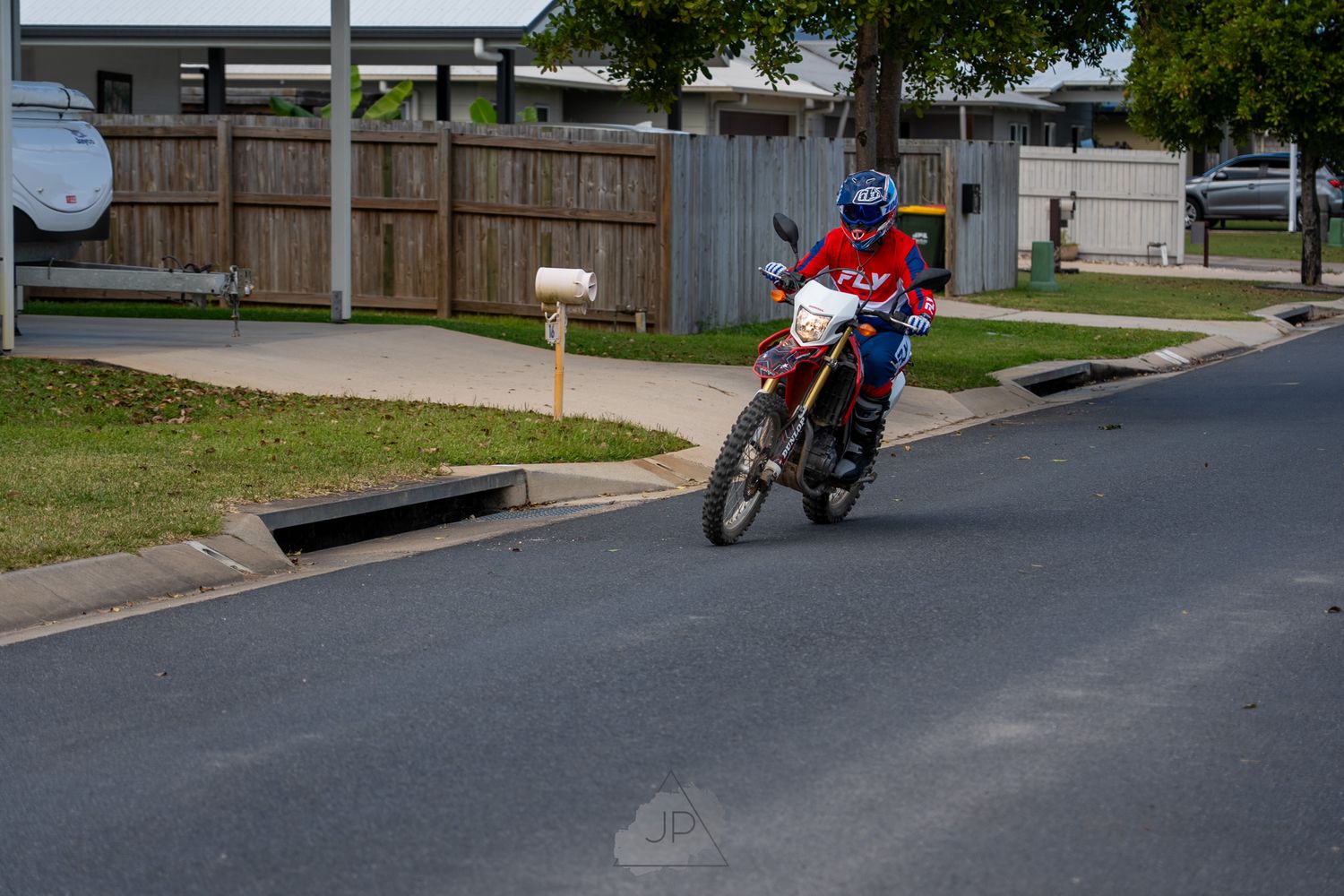 Motorcyclist rides down a residential street with green trees and wooden fences visible along the roadside.