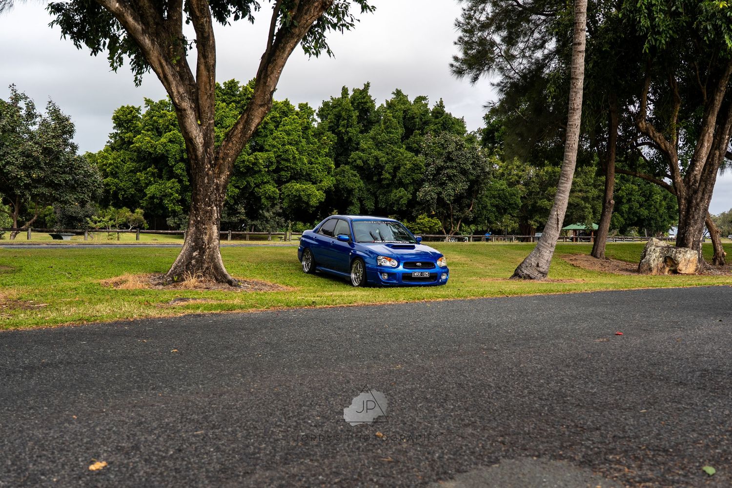 A bright blue Subaru WRX parked on a paved road under tall trees in a tropical setting.