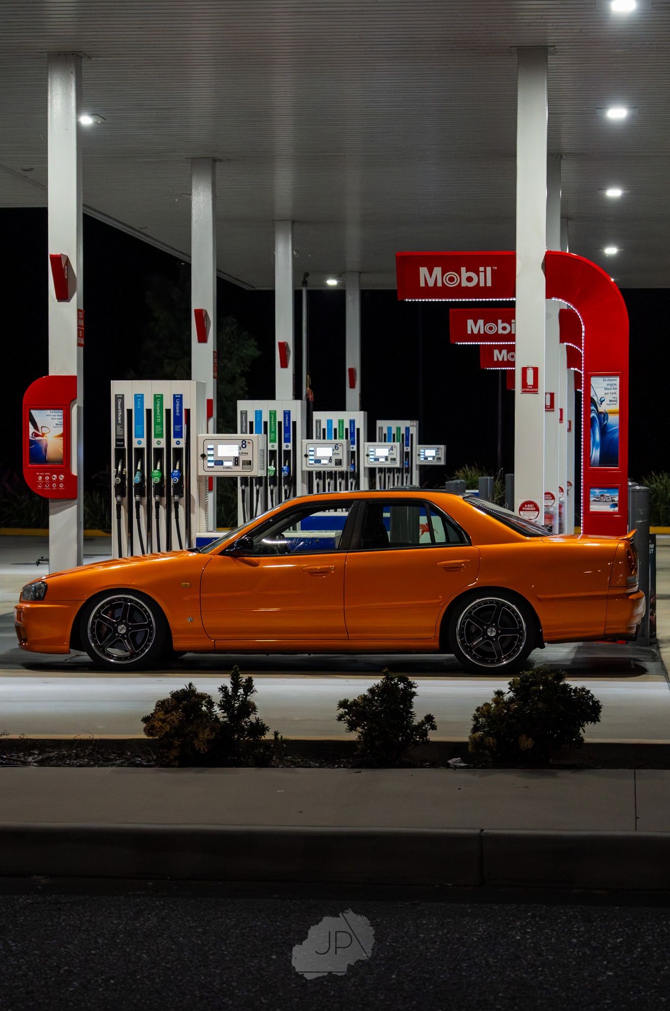 Orange sports car parked at illuminated Mobil gas station at night with fuel pumps visible in background.