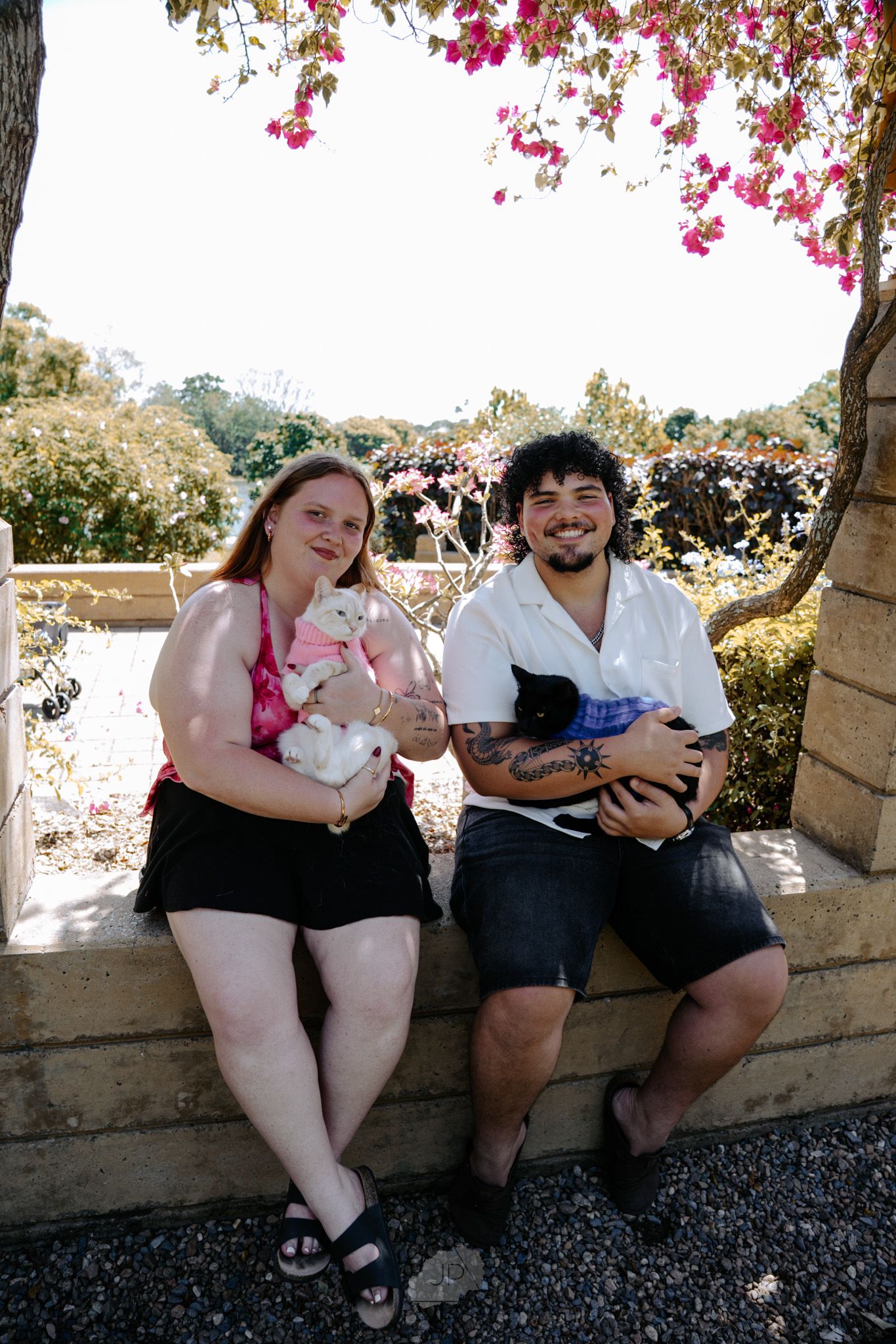 Three people sit together on a stone ledge holding small pets while pink flowers bloom overhead in a garden setting.