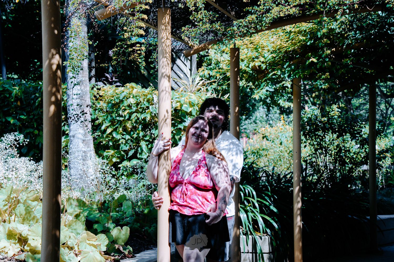 Two people walk through a lush garden path surrounded by tropical plants and tall trees.