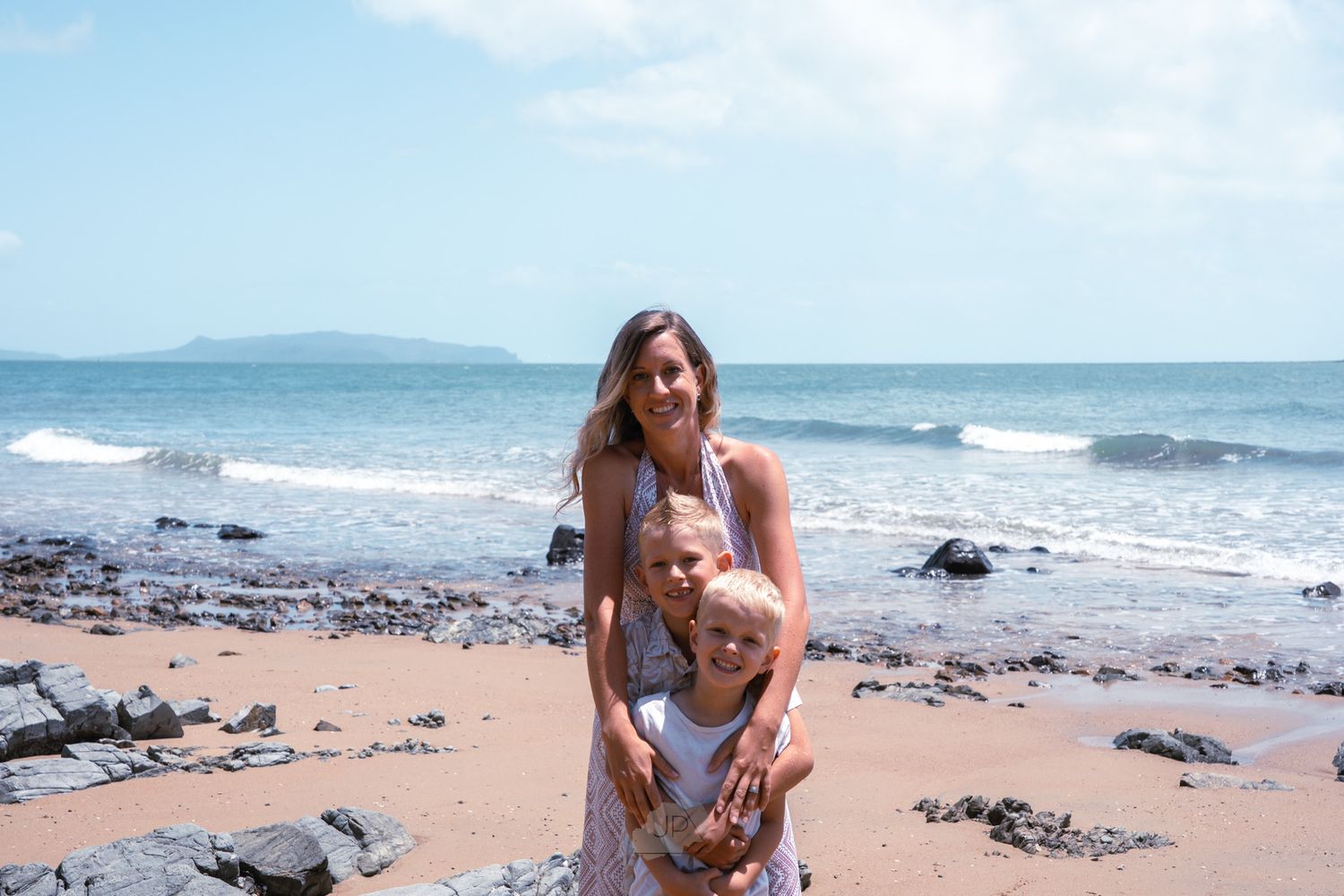 People enjoying a sunny day at a rocky beach with waves crashing against the shore and mountains in the distance.