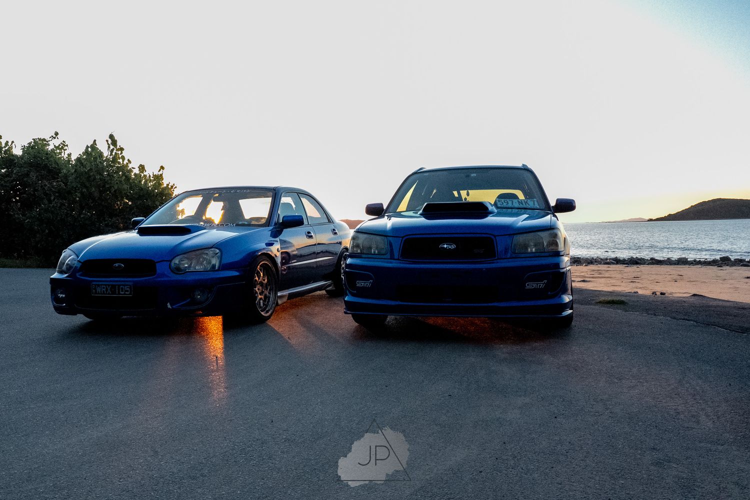 Blue Subaru WRX STI sports cars lined up at sunset with dramatic lighting and ocean backdrop.