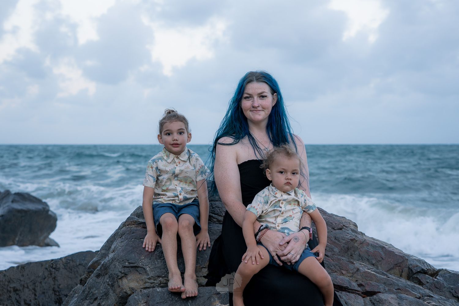 A family sits together on rocky coastal outcrop with crashing waves and cloudy sky in the background.