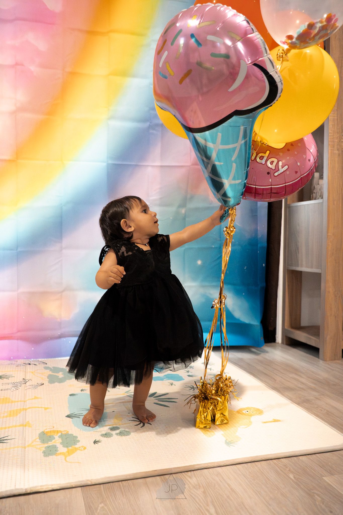 Colorful balloons float against a pink and orange gradient backdrop in a room with hardwood floors and gold accents.