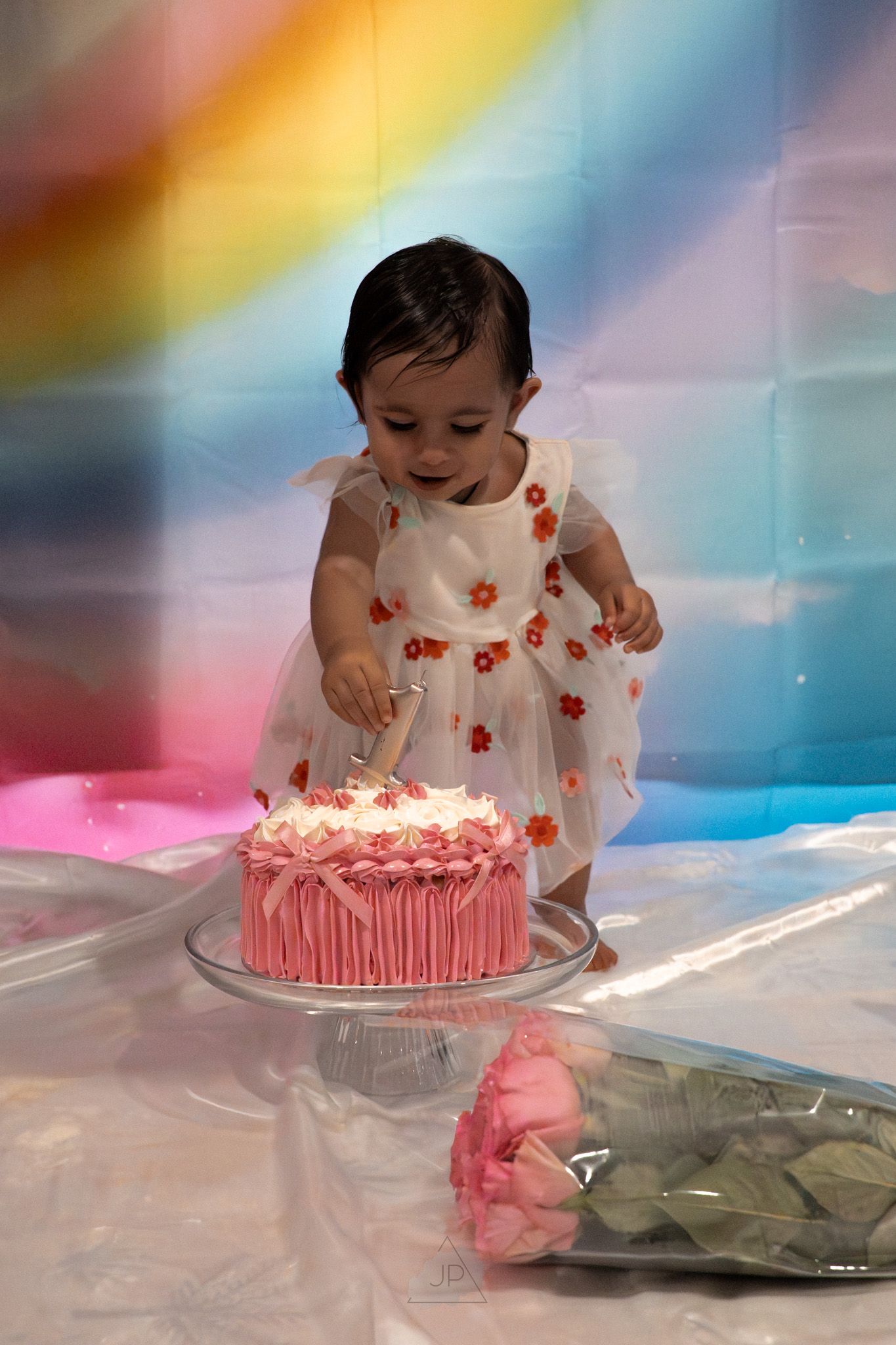 A small child in a floral dress leans over a pink birthday cake with lit candles on a colorful rainbow background.