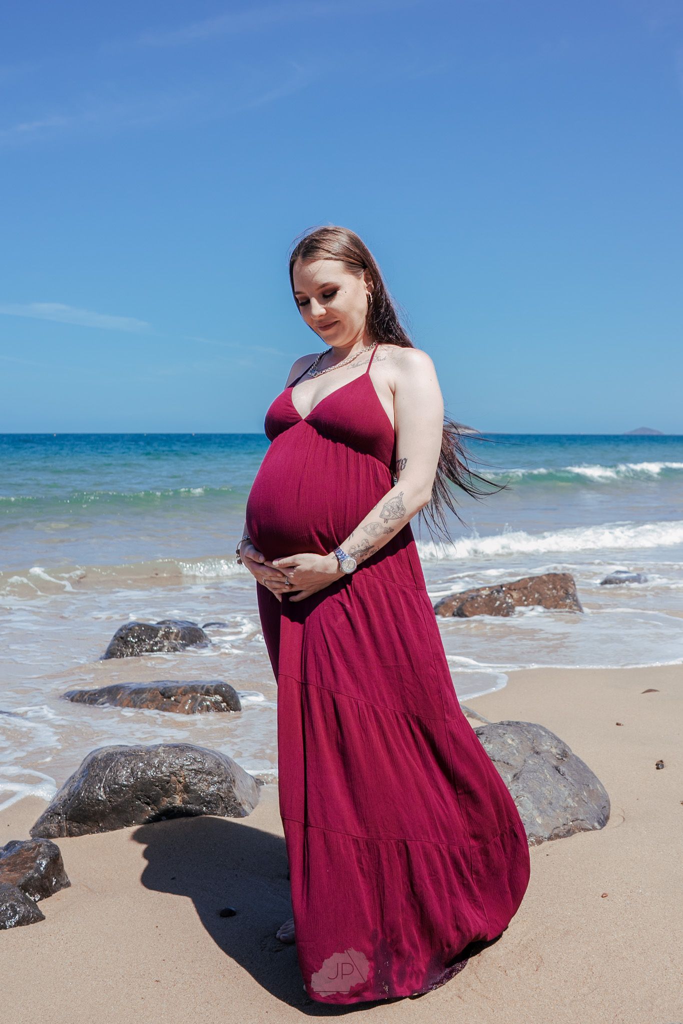 A person in a flowing burgundy dress poses gracefully on a sandy beach with ocean waves and rocks in the background.