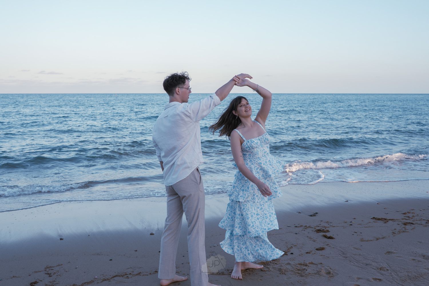 A couple dances together on a beach at sunset with ocean waves in the background.