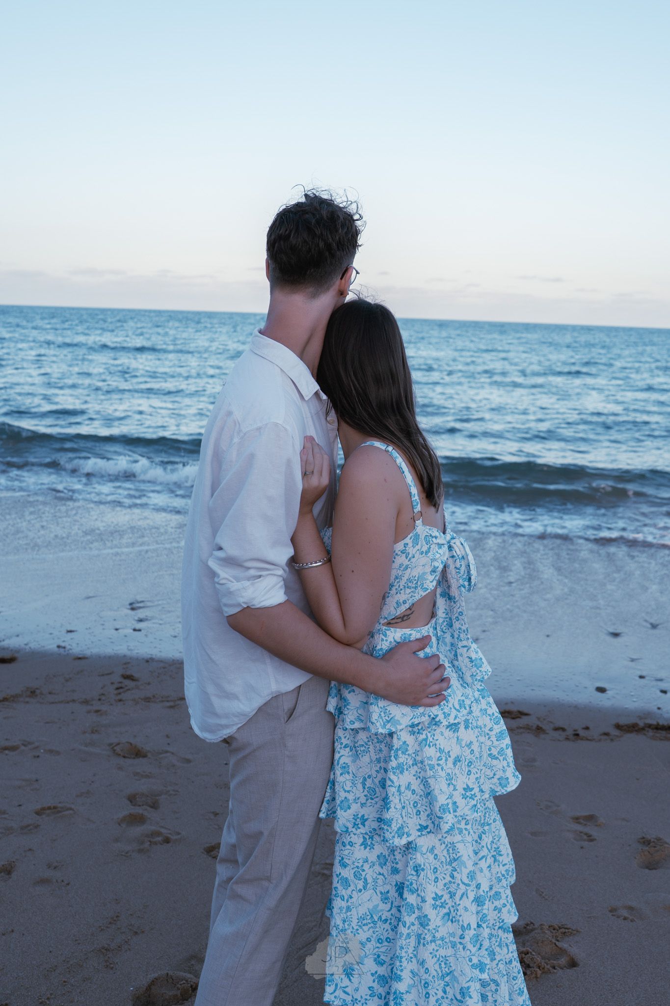 A couple in white and blue floral attire share a romantic moment on a beach at sunset with ocean waves in the background.