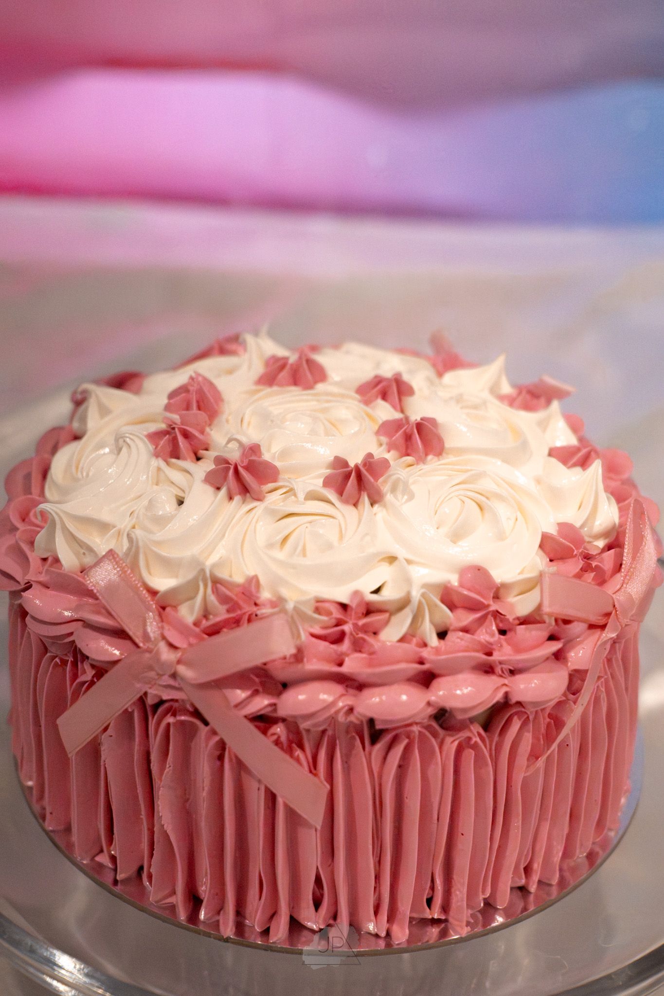Pink and white buttercream frosted cake with rosettes and ruffled sides on a silver cake stand.