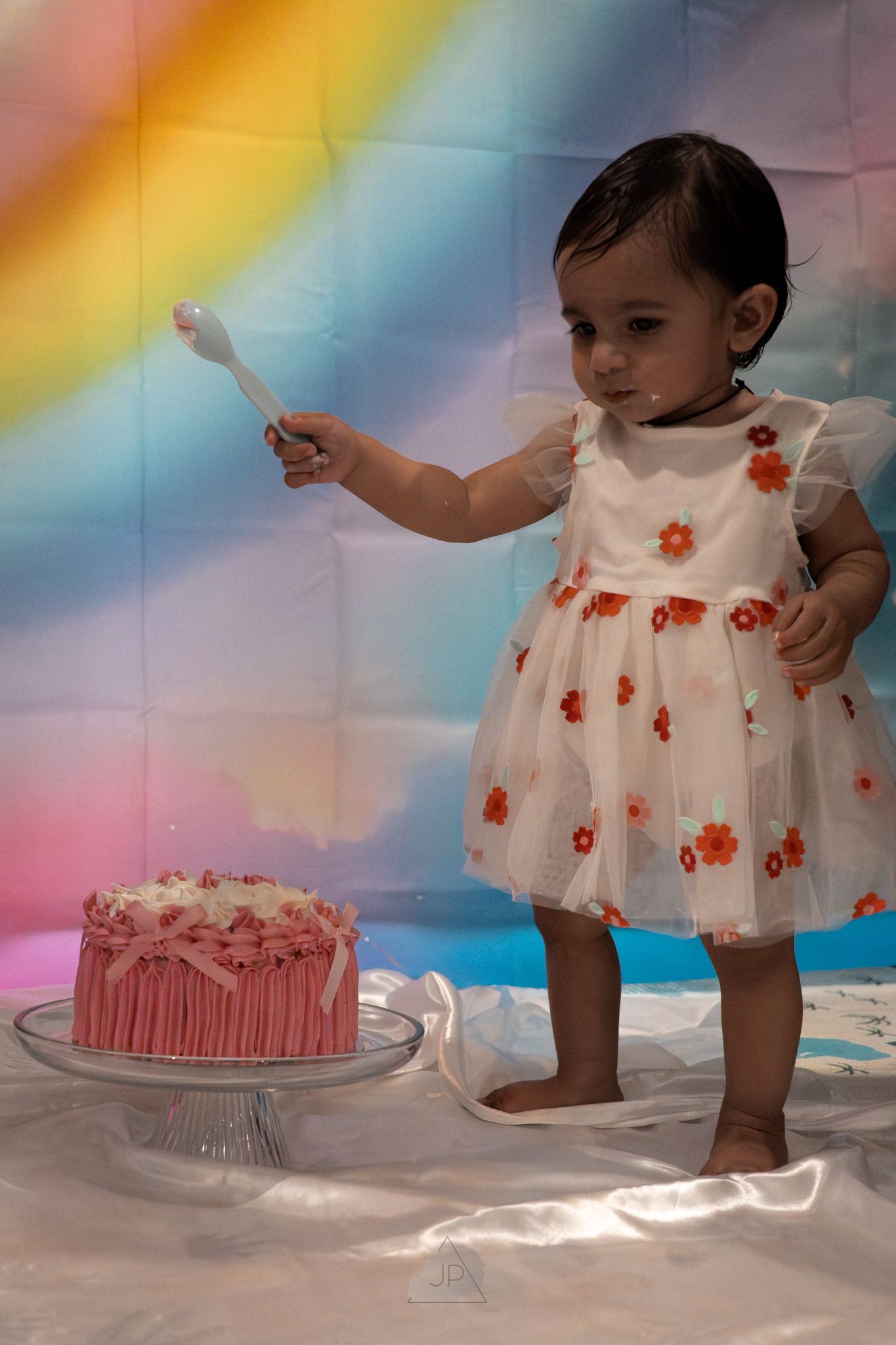 A small child in a floral dress reaches toward a pink birthday cake while standing against a rainbow-colored backdrop.