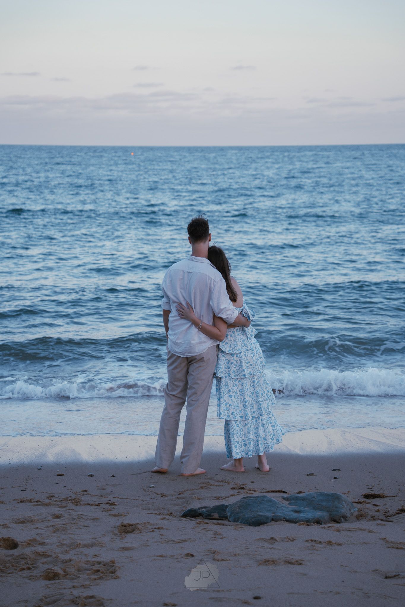 A couple in white attire stands together watching a sea turtle on a sandy beach at sunset.