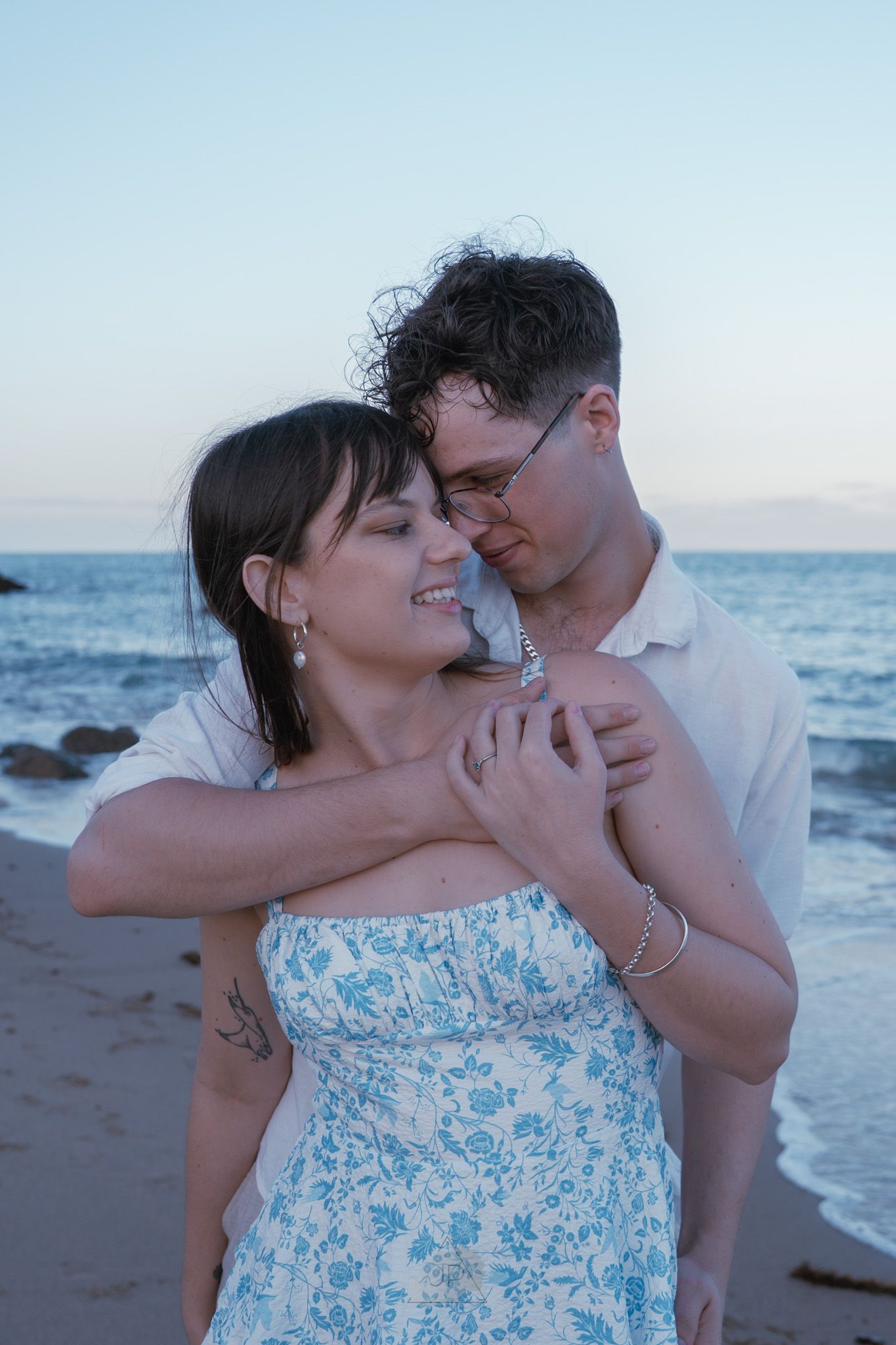 A romantic couple embraces on a beach at sunset, with the woman wearing a blue and white patterned sundress.