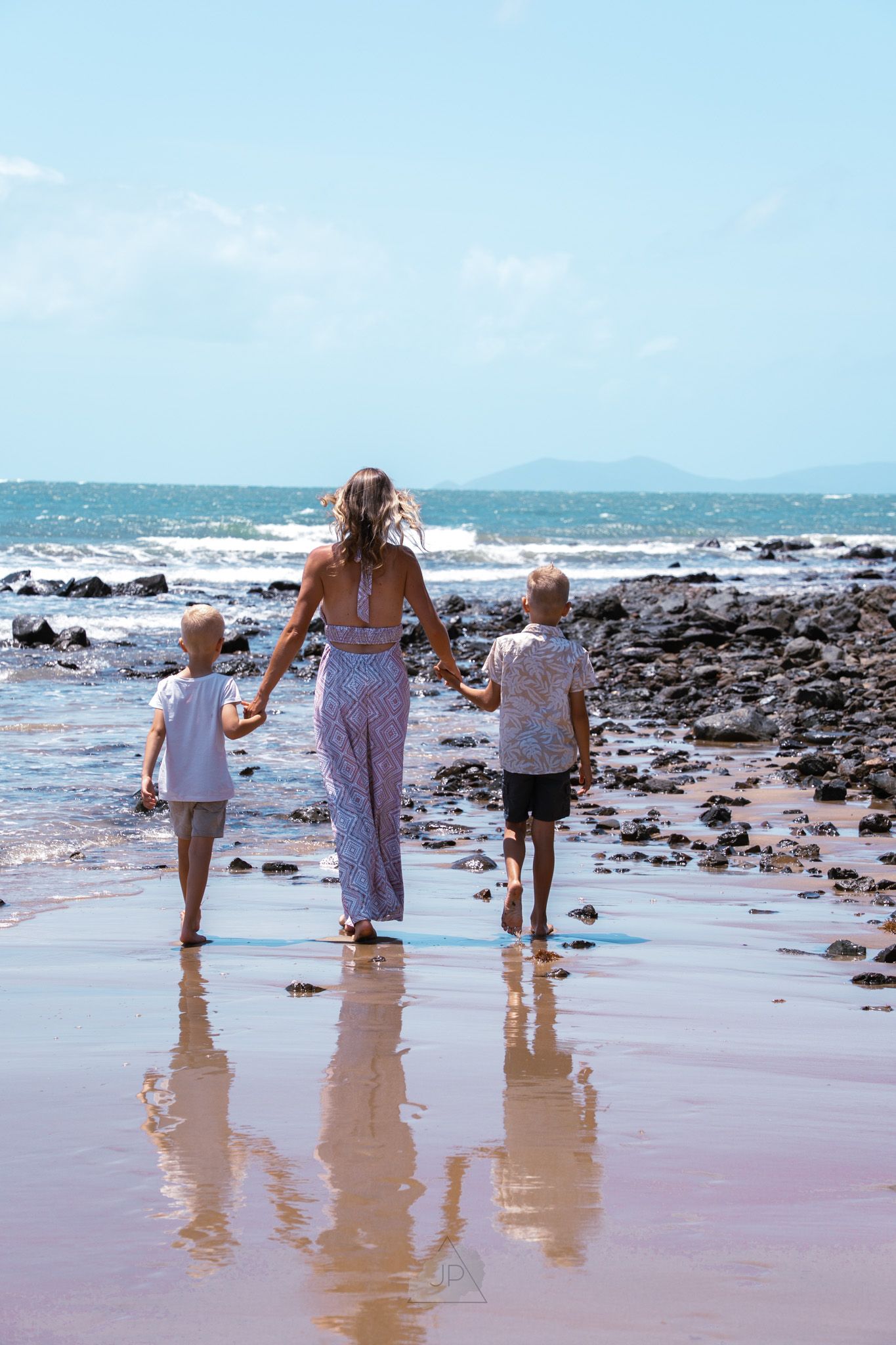 Three figures walk hand in hand along a rocky beach shoreline with waves crashing in the background.