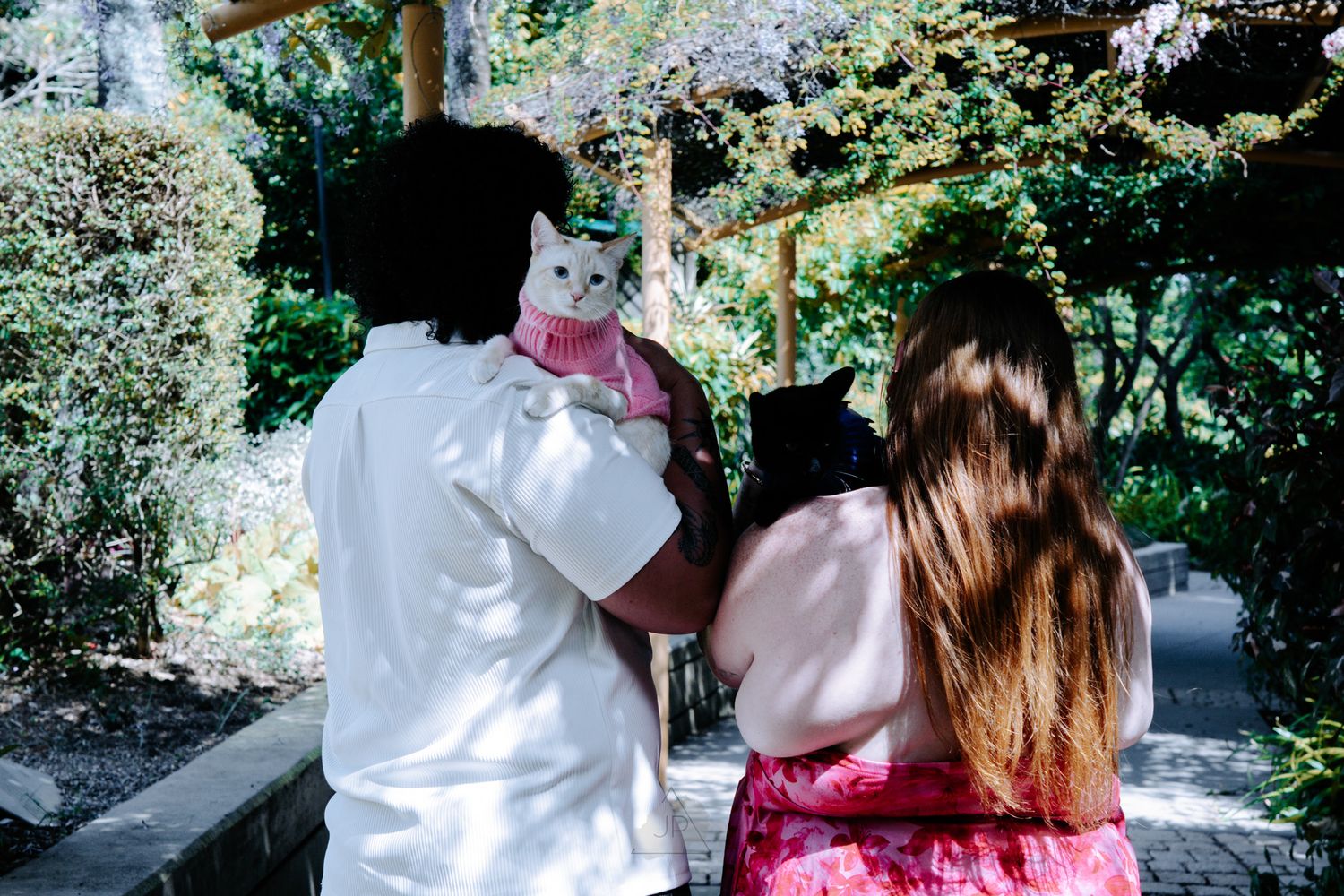 Two people in pink and white outfits sit together on a bench in a lush garden setting.