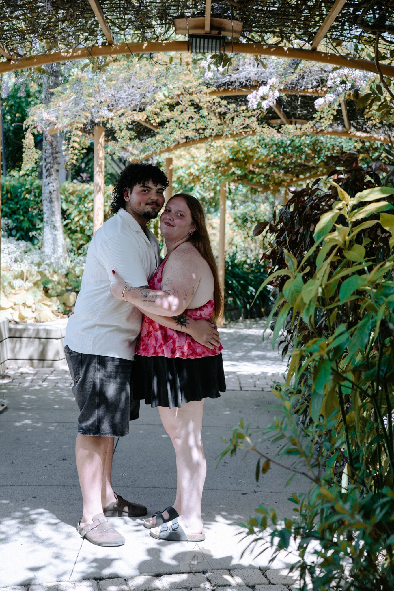 A couple dances together in a garden setting under dappled sunlight and greenery.