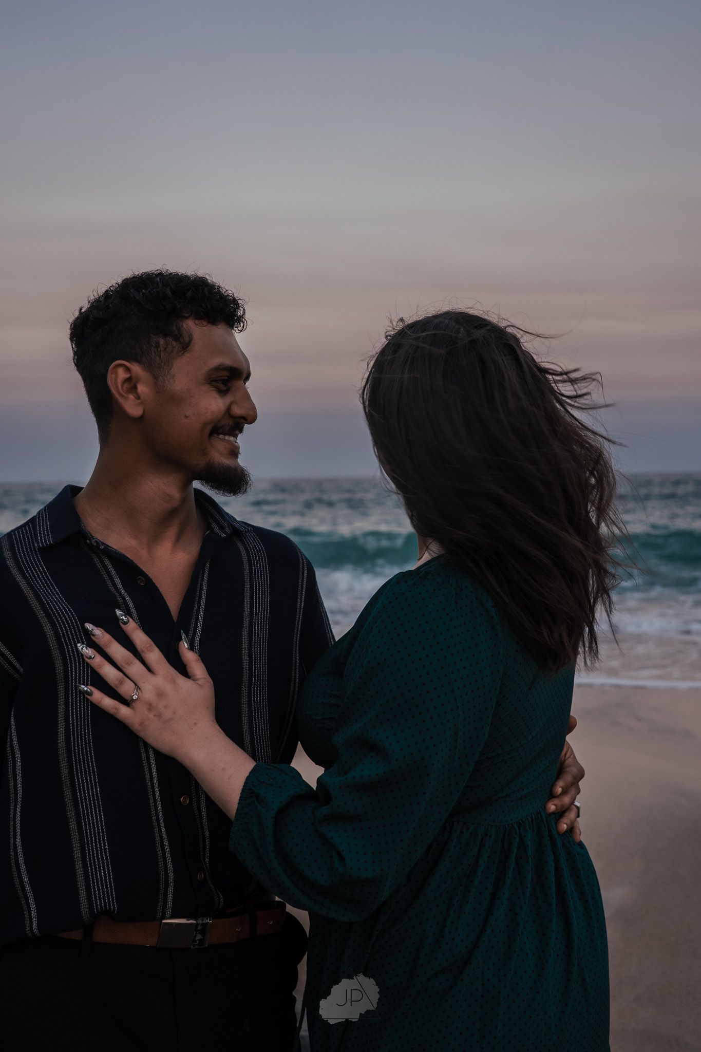 A couple embraces on a moody beach at sunset with waves crashing in the background.
