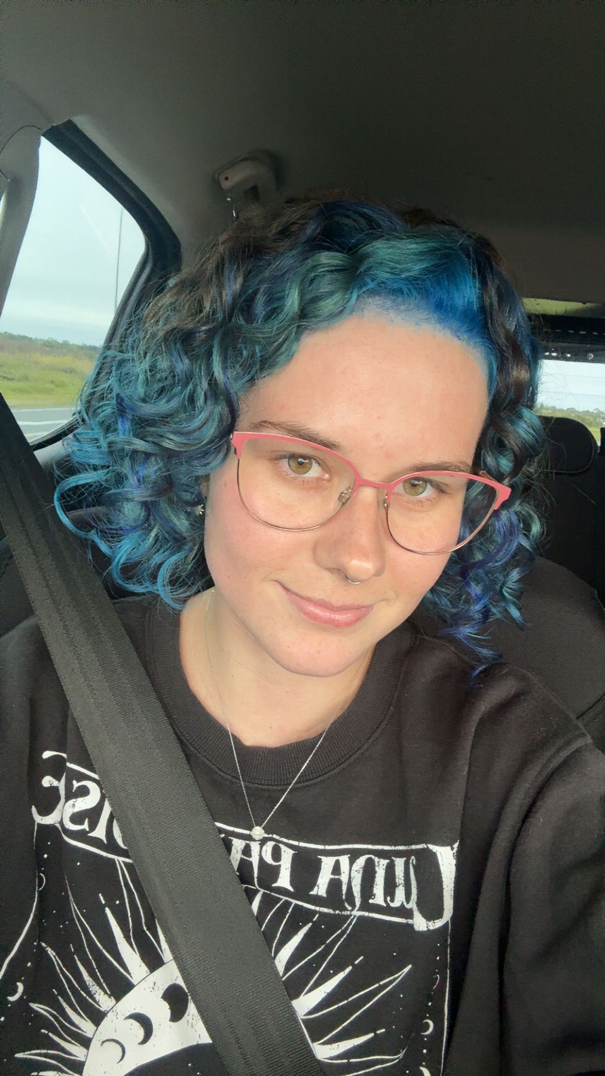 Someone with vibrant blue curly hair takes selfies wearing a black graphic t-shirt inside a car.