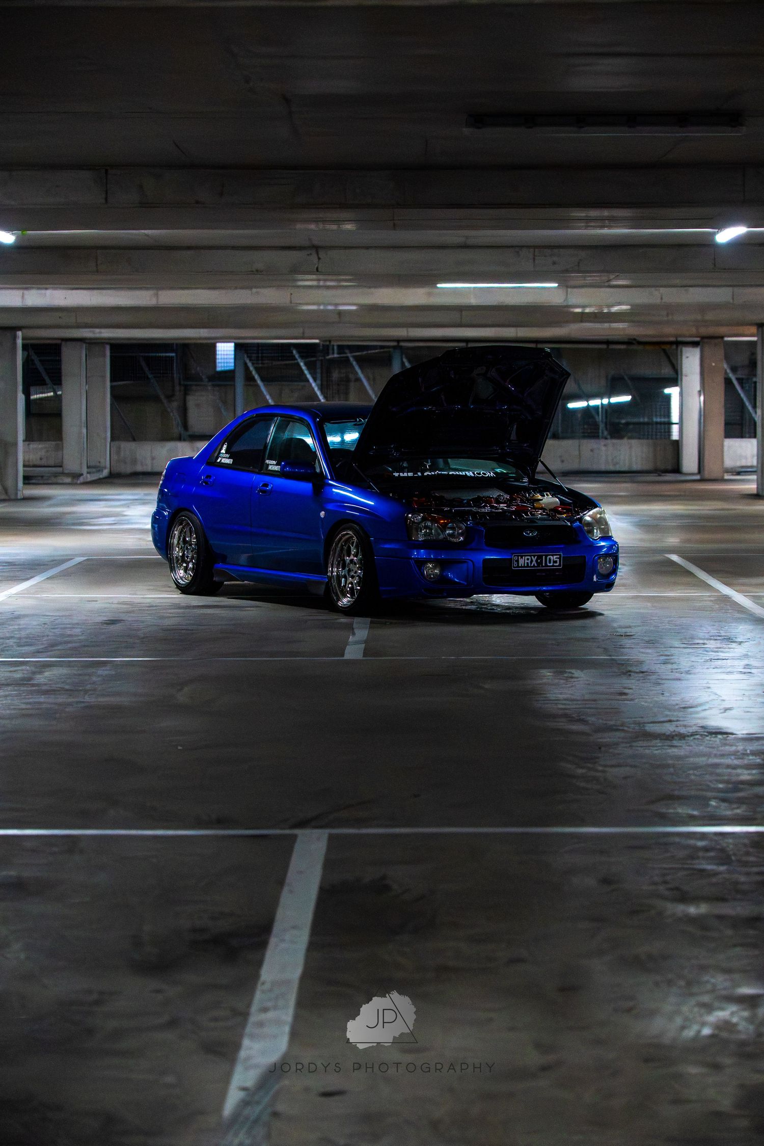 Blue modified sports car parked in dimly lit concrete parking garage from multiple angles showing sleek customizations.