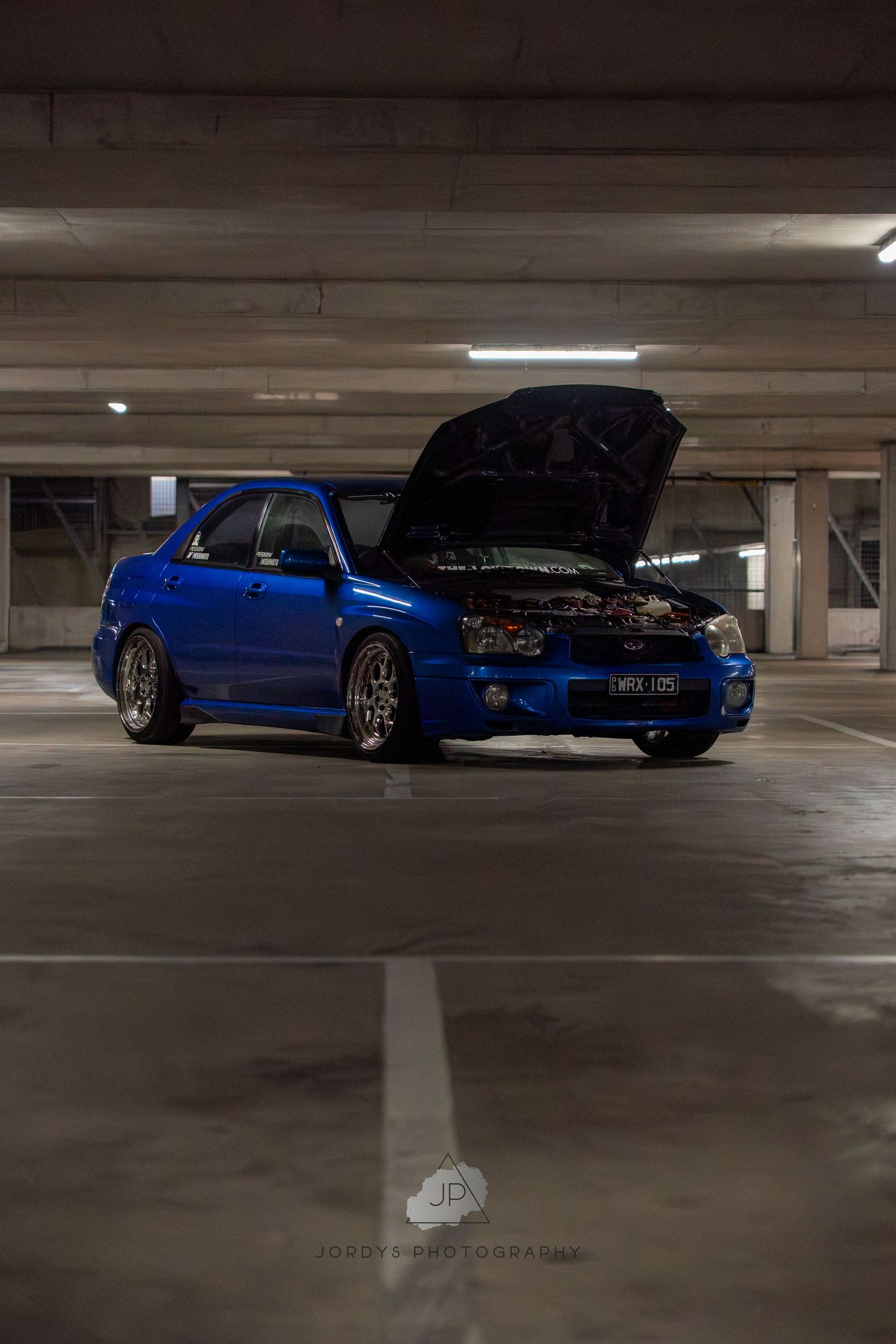 A dark blue modified sports car parked in a dimly lit indoor parking garage from multiple angles.