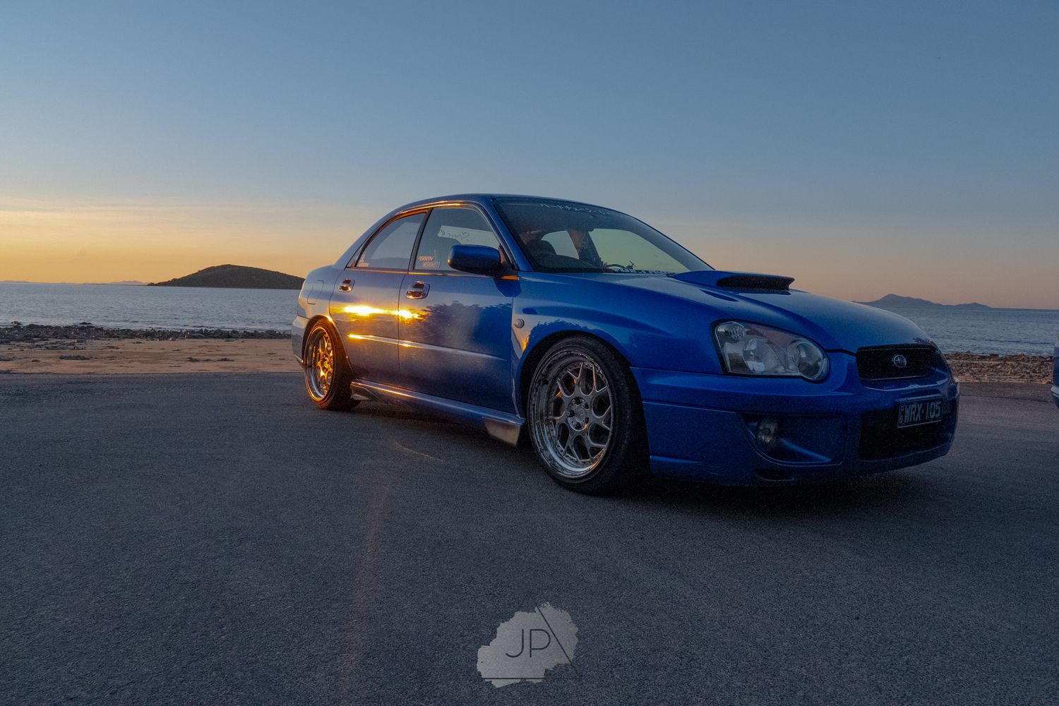 Blue Subaru WRX STI parked on a beach at sunset with its lights on against a colorful evening sky.