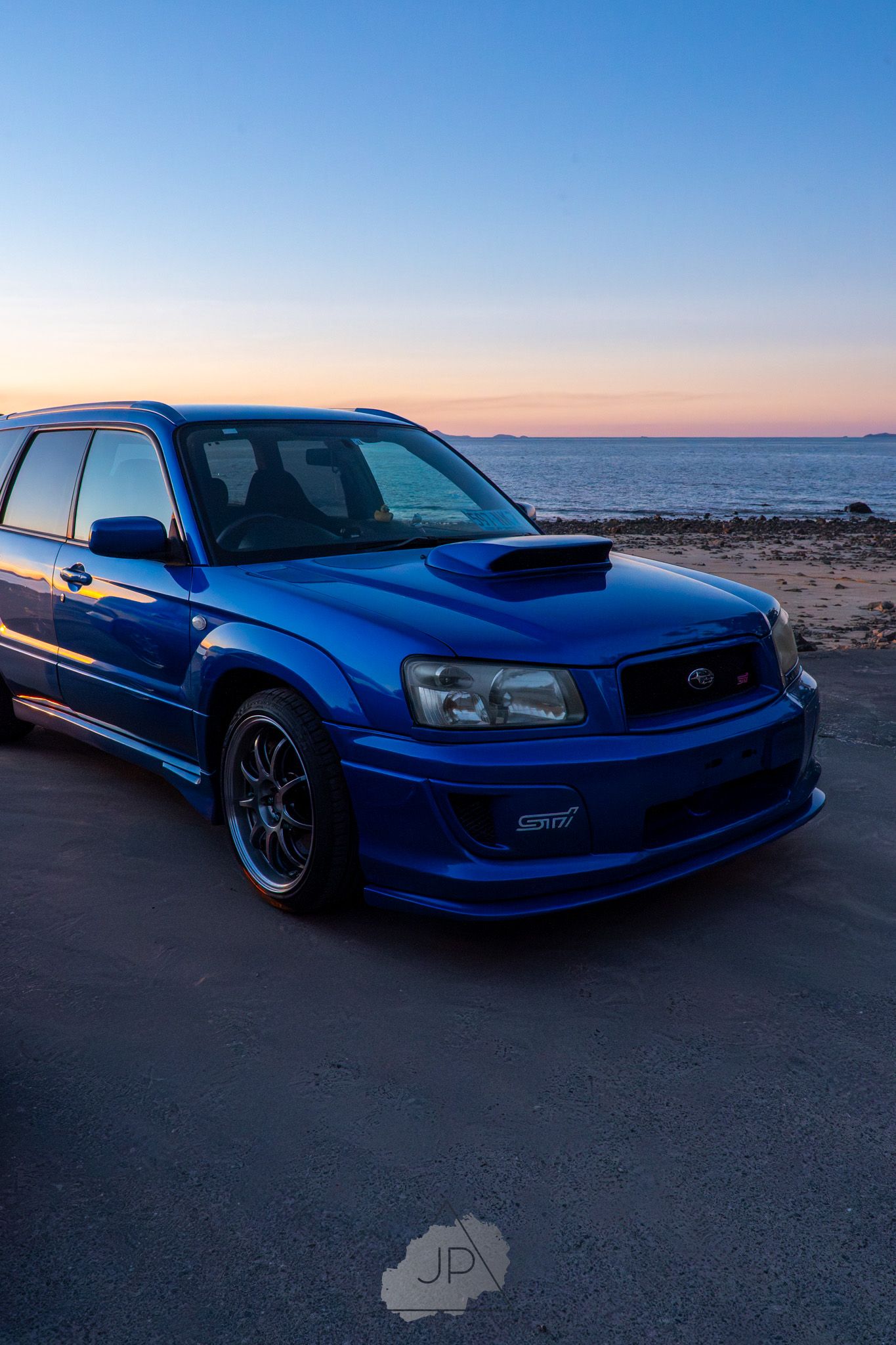 Blue modified sports car photographed at sunset against desert landscape and sky backdrop.