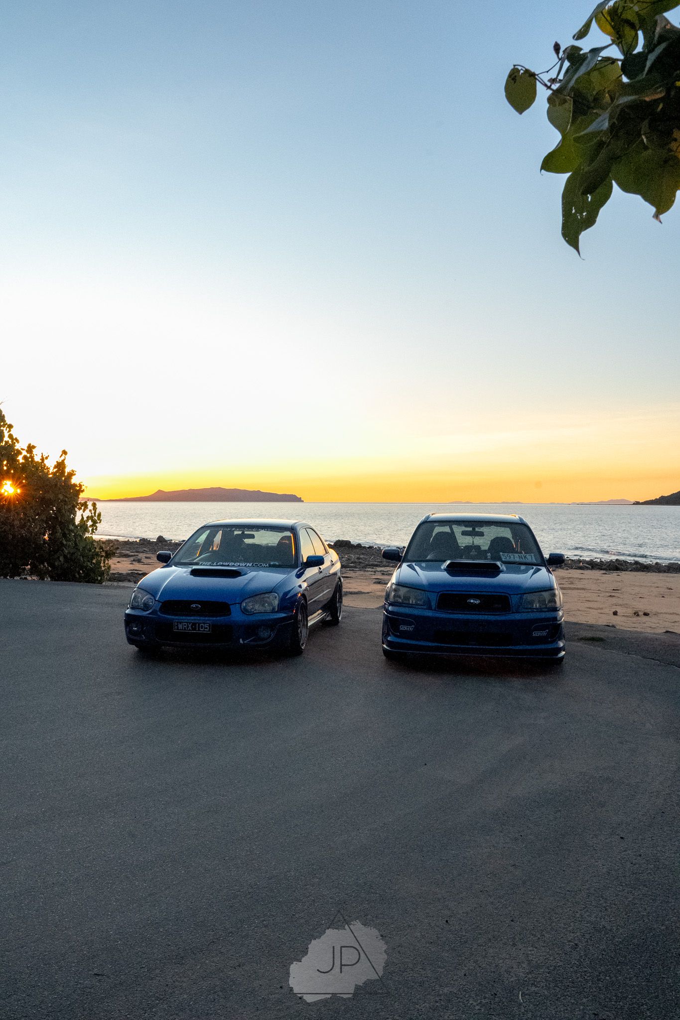 Two blue sports cars parked at a beach during sunset with orange skies and mountains in the distance.
