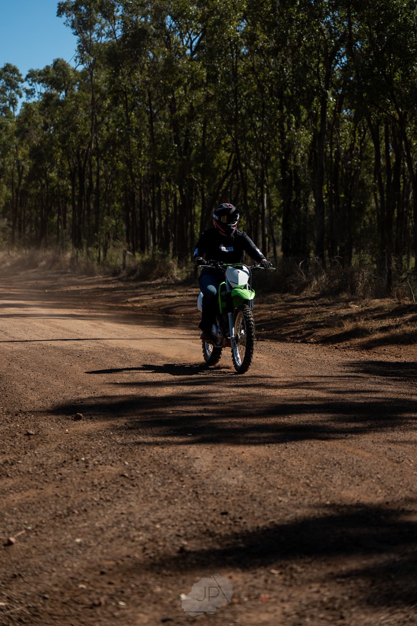 A motorcyclist in bright gear rides along a rugged outback trail surrounded by gum trees.