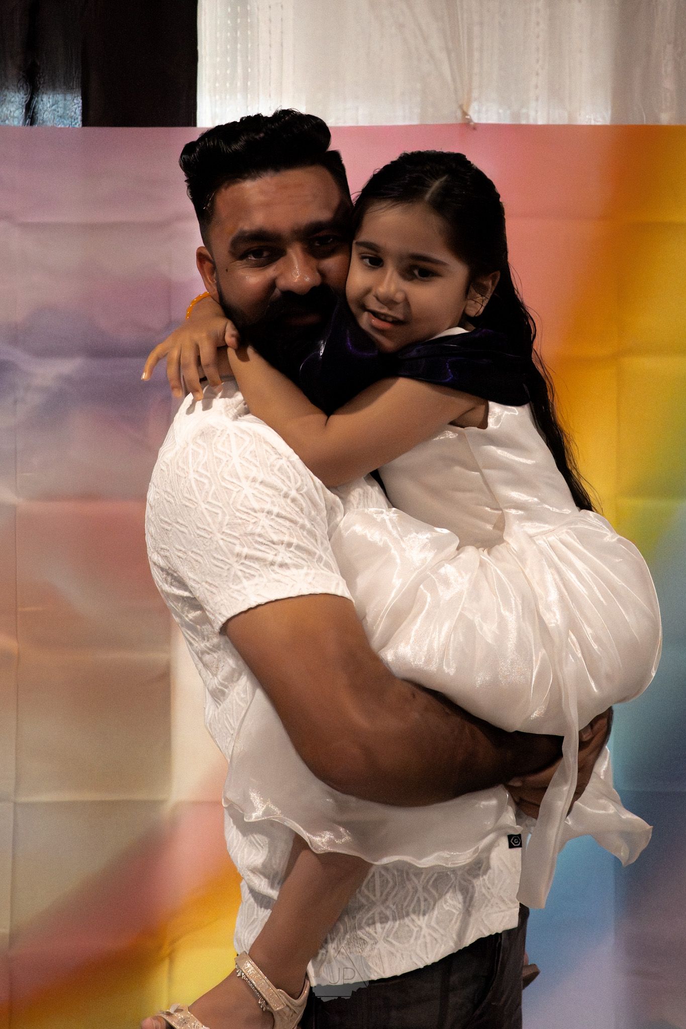 A parent and child wearing white outfits embrace in a warm hug against a colorful background.