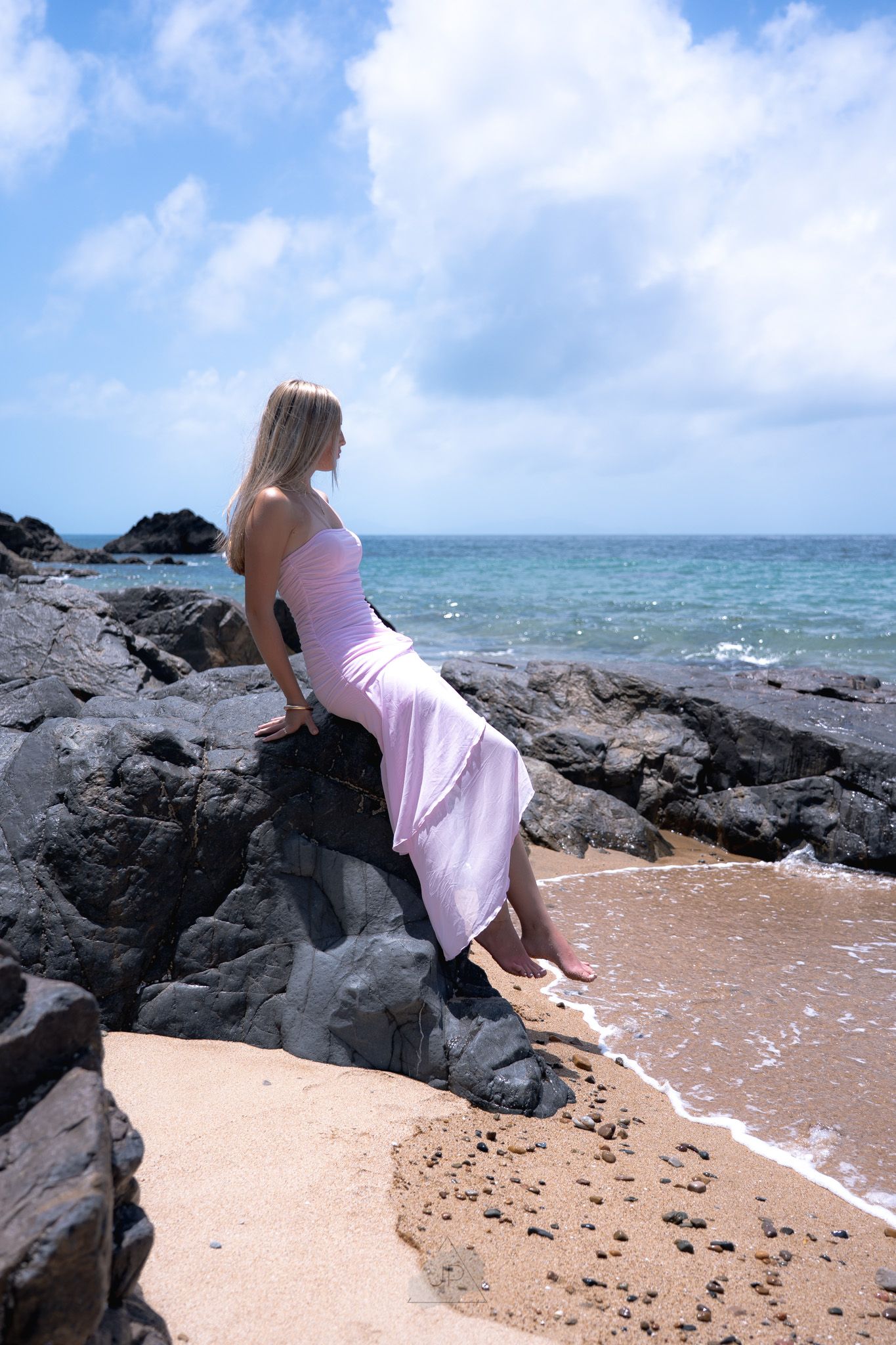 A person in a pink dress sits on dark volcanic rocks at the edge of a beach where waves meet the shore.