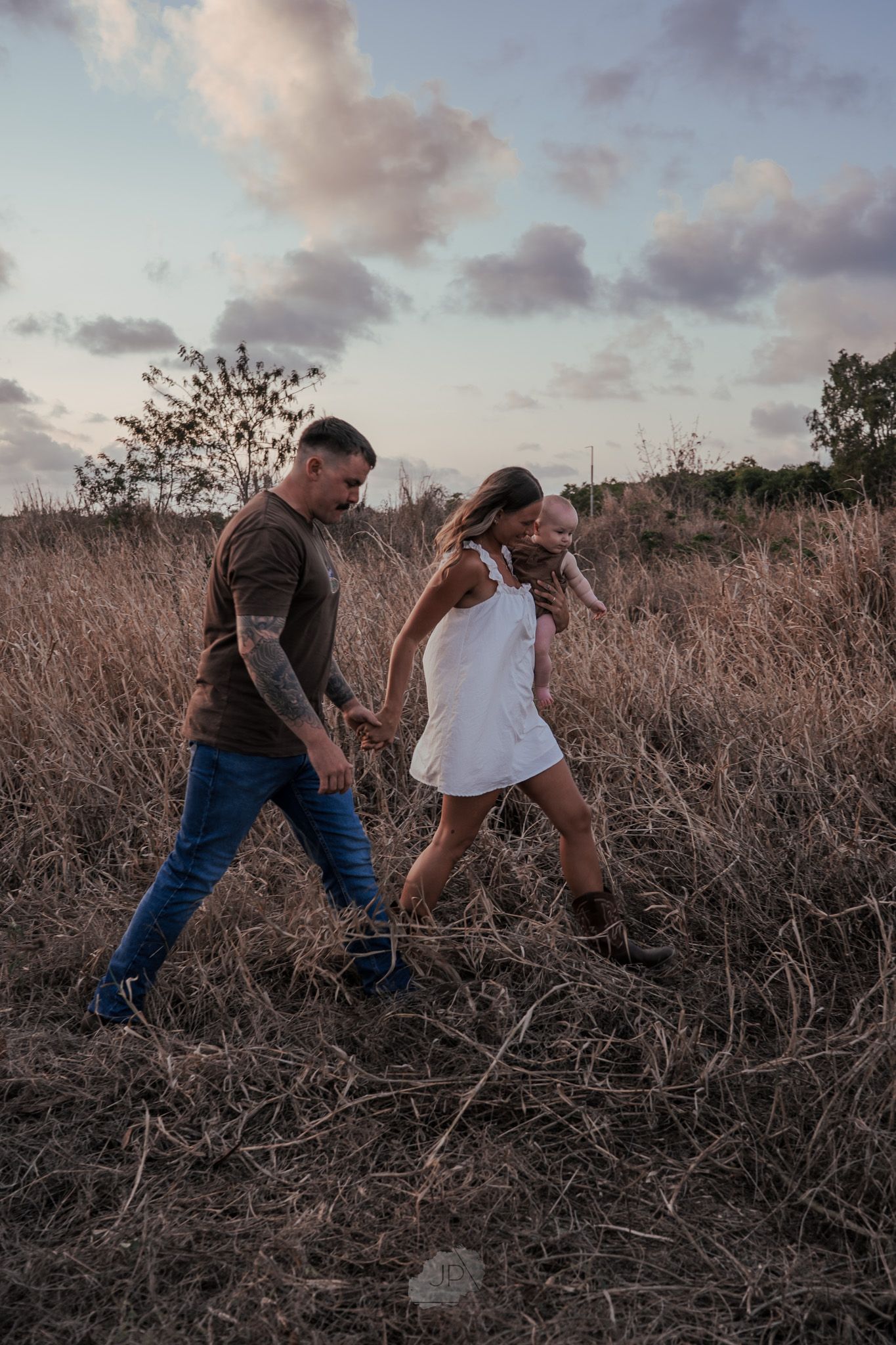 A couple playfully interacts in a field of dry brush during sunset with dramatic cloudy skies overhead.