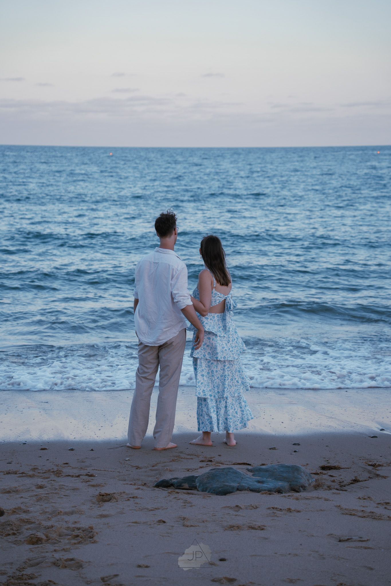 A romantic beach scene shows two people standing together by the ocean during a tranquil evening.