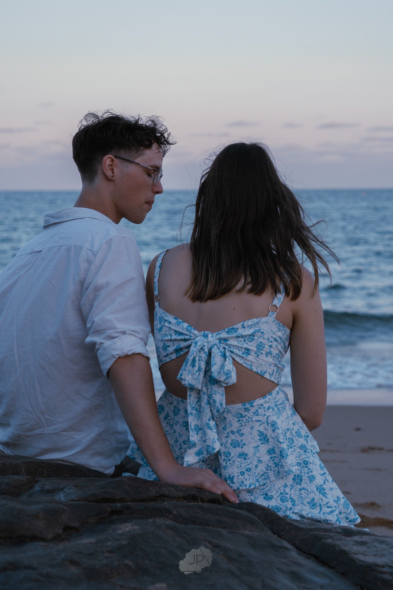 A couple sits together on a beach at sunset wearing white and blue floral clothing while looking at the ocean.