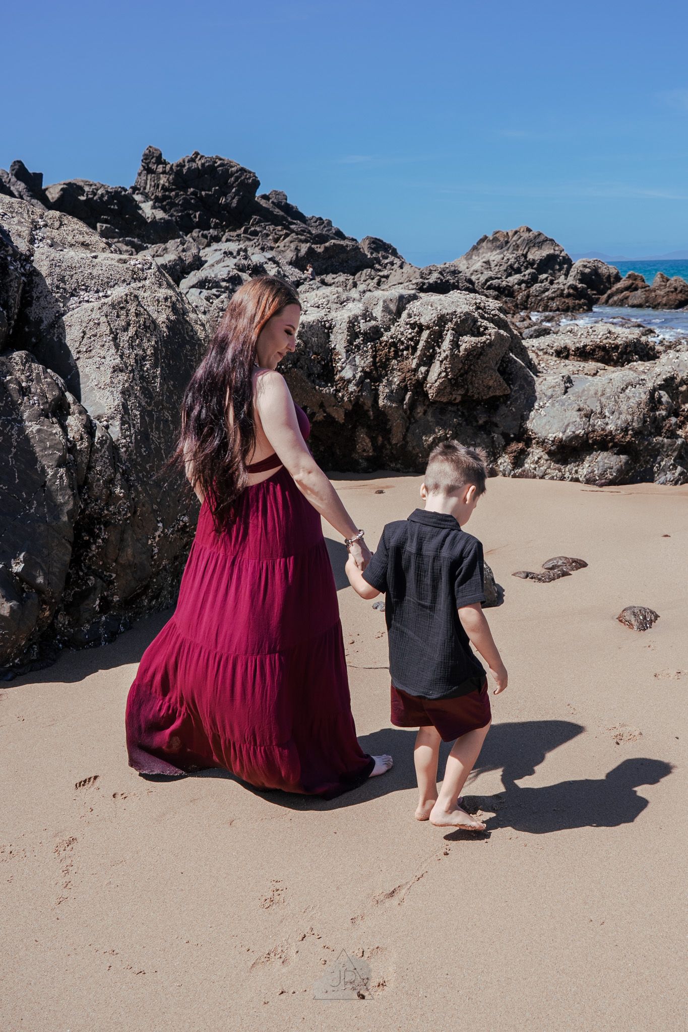 Two people explore a rocky beach with turquoise waters and volcanic formations on a sunny day.