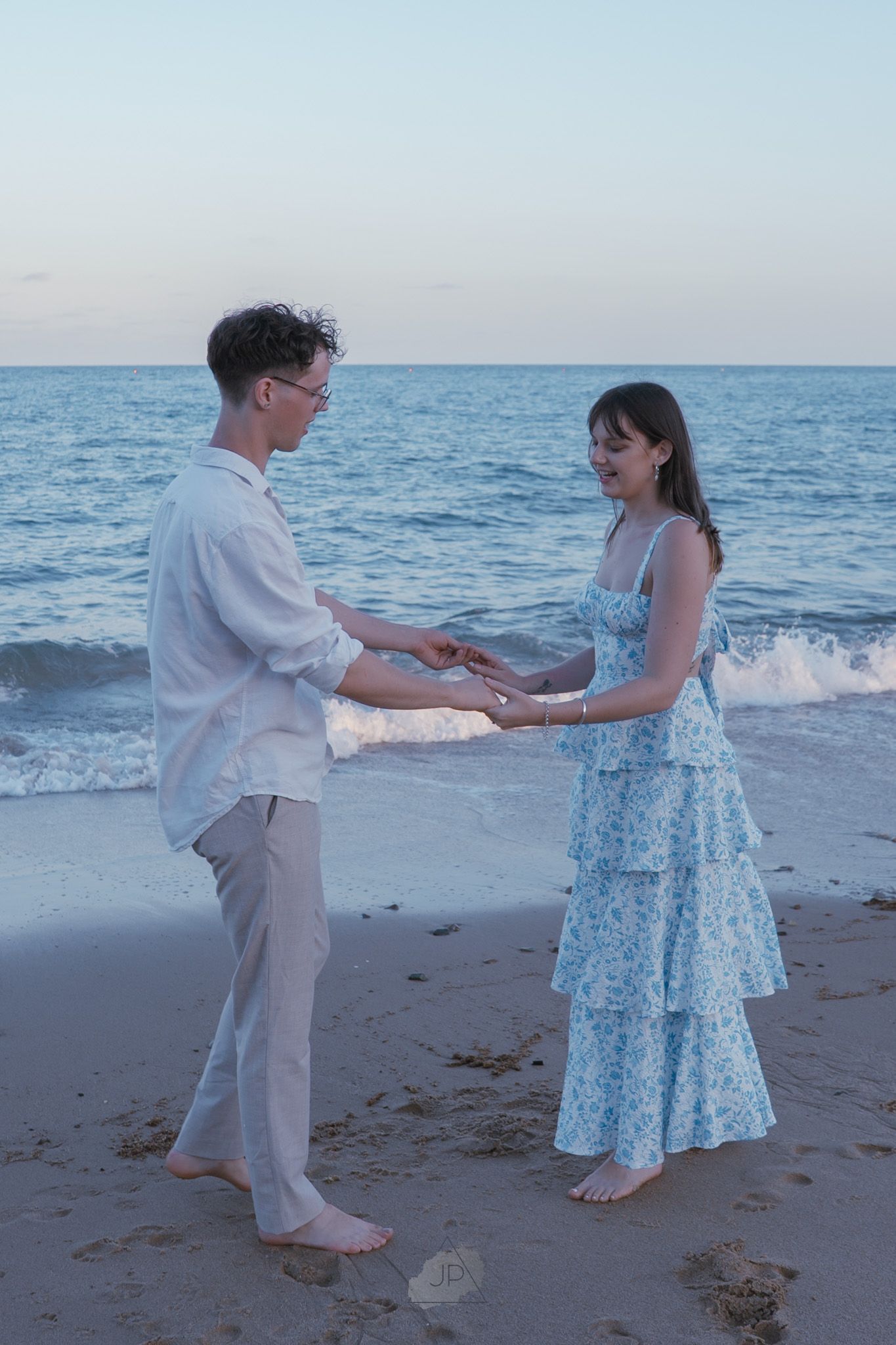 A couple in white and blue outfits dancing together on a beach at sunset with ocean waves in the background.