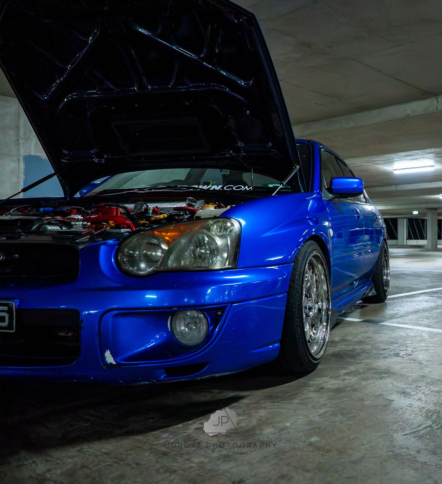 Bright blue Subaru Impreza WRX with open hood parked in dimly lit garage showing performance modifications.