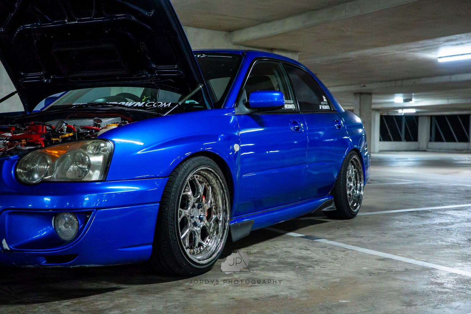 Blue Subaru WRX with custom wheels and body modifications sits in a parking garage under fluorescent lighting.