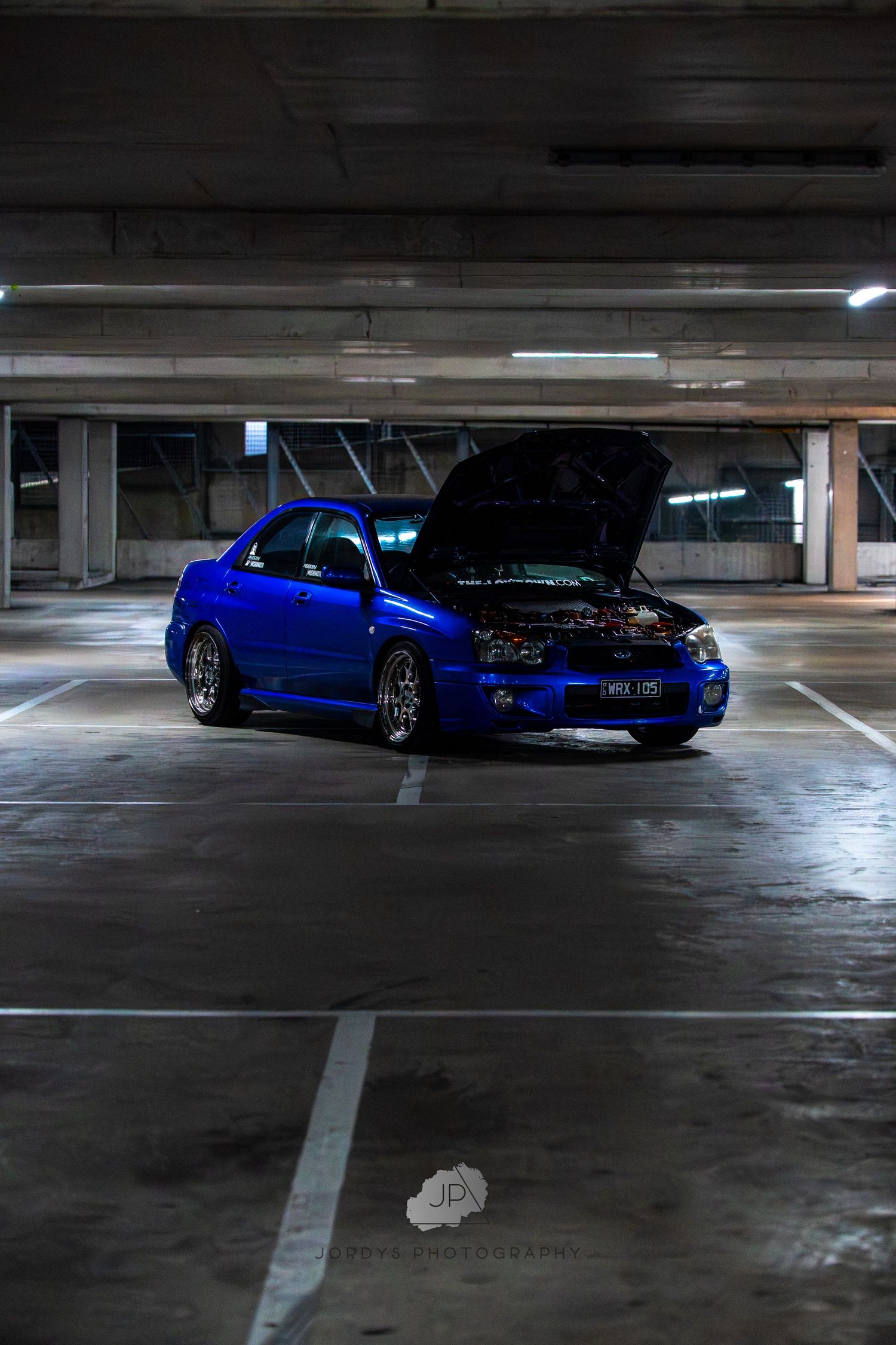 A modified blue Subaru WRX is parked in a dimly lit underground parking garage at night.
