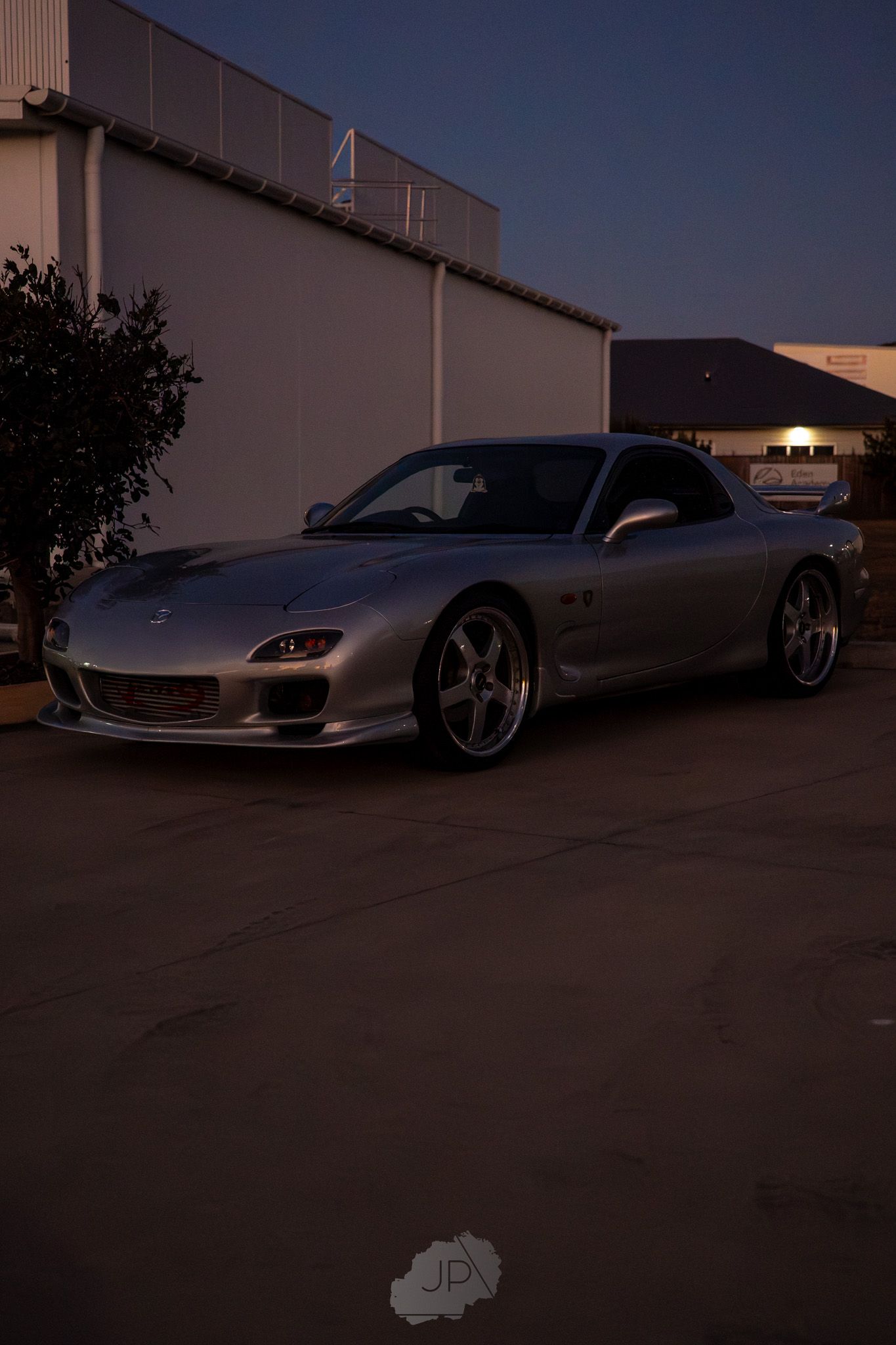 A silver Mazda RX-7 sports car parked at dusk with custom wheels against a building exterior.