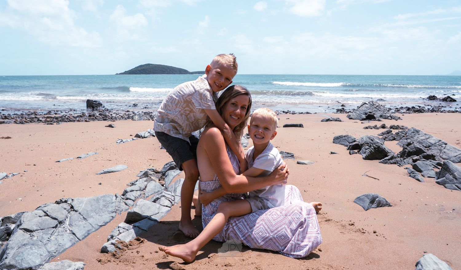 A family enjoys a sunny day together on a rocky beach with an island visible in the ocean background.